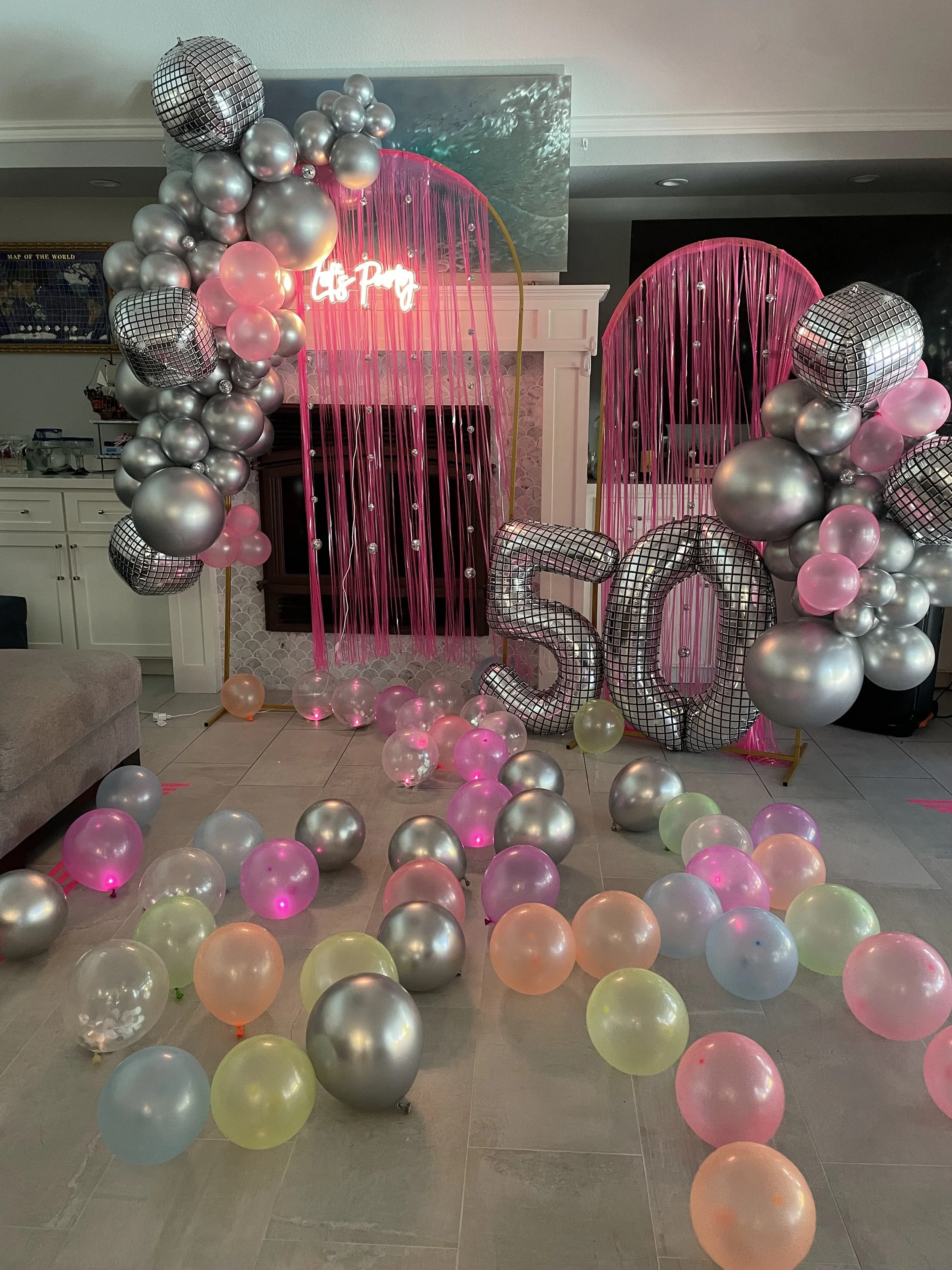 Decorative party backdrop with pink curtains, large silver and black balloons, some with disco ball patterns, and a neon sign that reads 'It's Party time'. There are also multicolored balloons scattered on the floor. 50th birthday celebration
