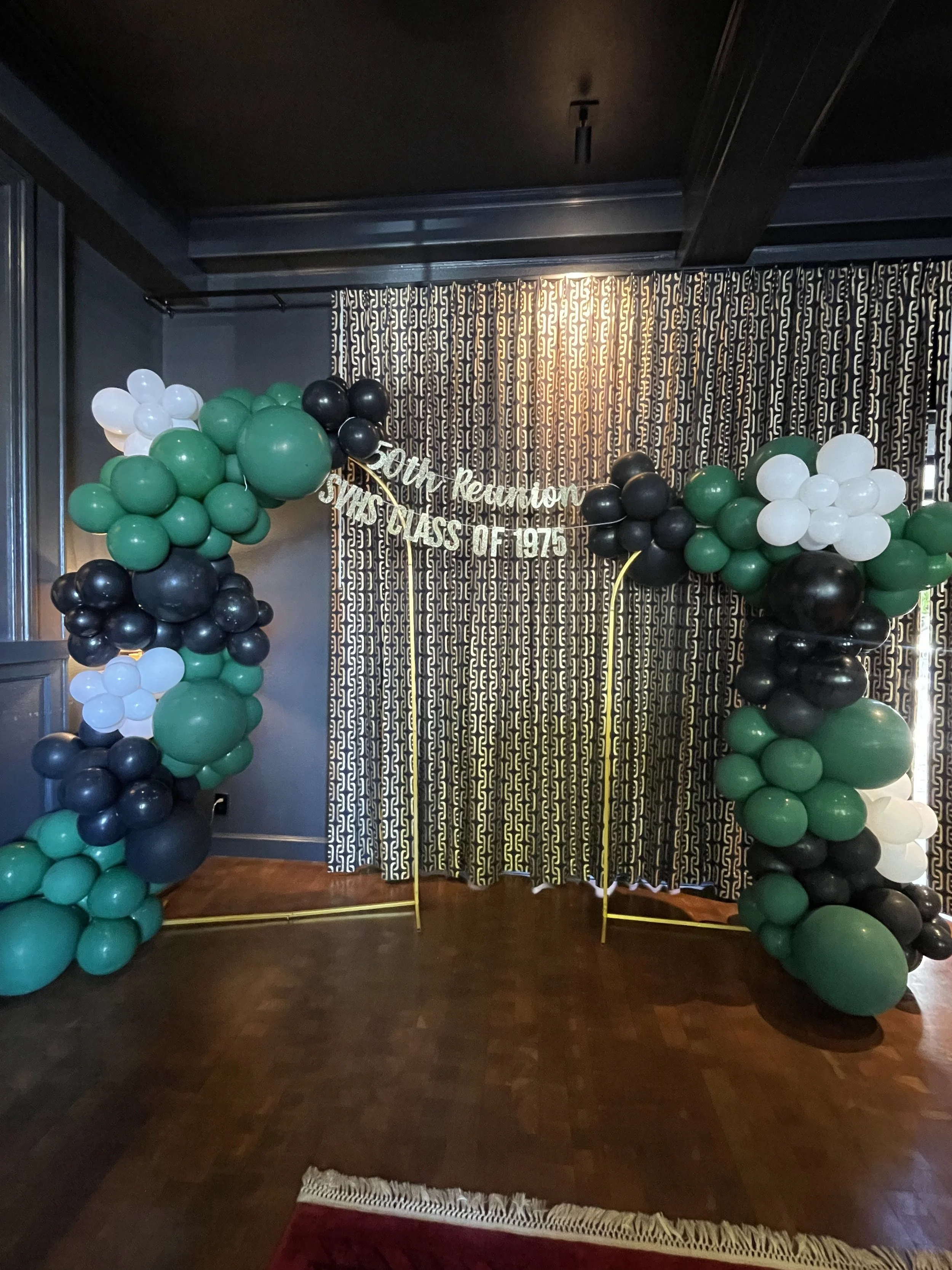 Balloon arch with green, black, and white balloons, and a banner that reads 'SVHS Class of 1975' in front of a black and gold patterned curtain. 50th Highschool reunion