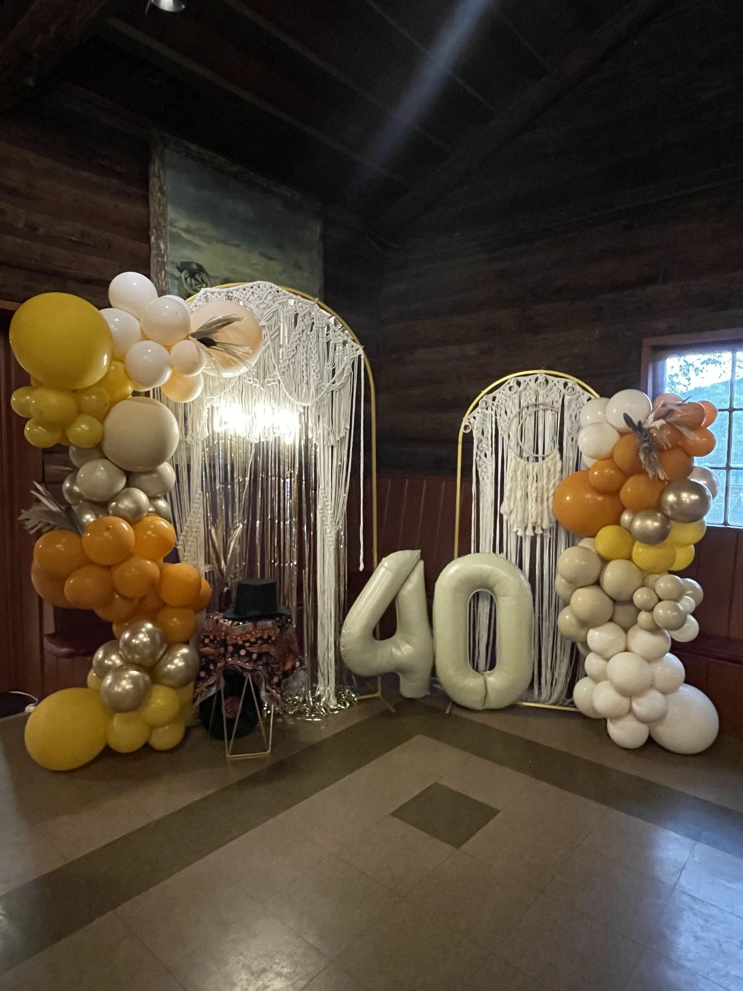 Decorative birthday balloon arch featuring black, white, gold, and transparent balloons with confetti, shaped balloons like a beer mug and a champagne bottle, large black number four balloons, and smaller balloons with messages like "Cheers to 40" and "Aged to Perfection."