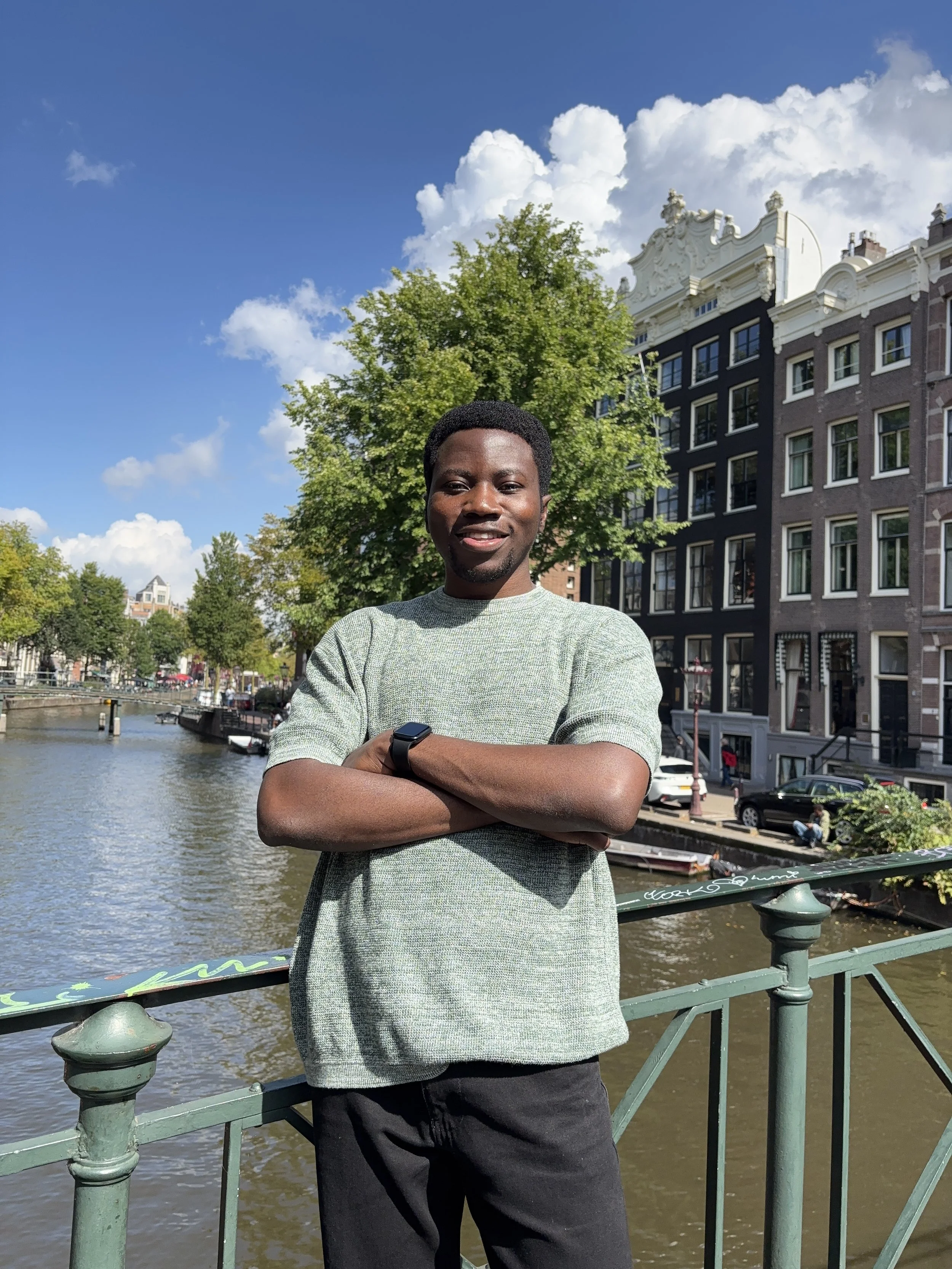 Young man with crossed arms standing in front of a canal with boats, trees, and historic buildings under a partly cloudy sky in Amsterdam.
