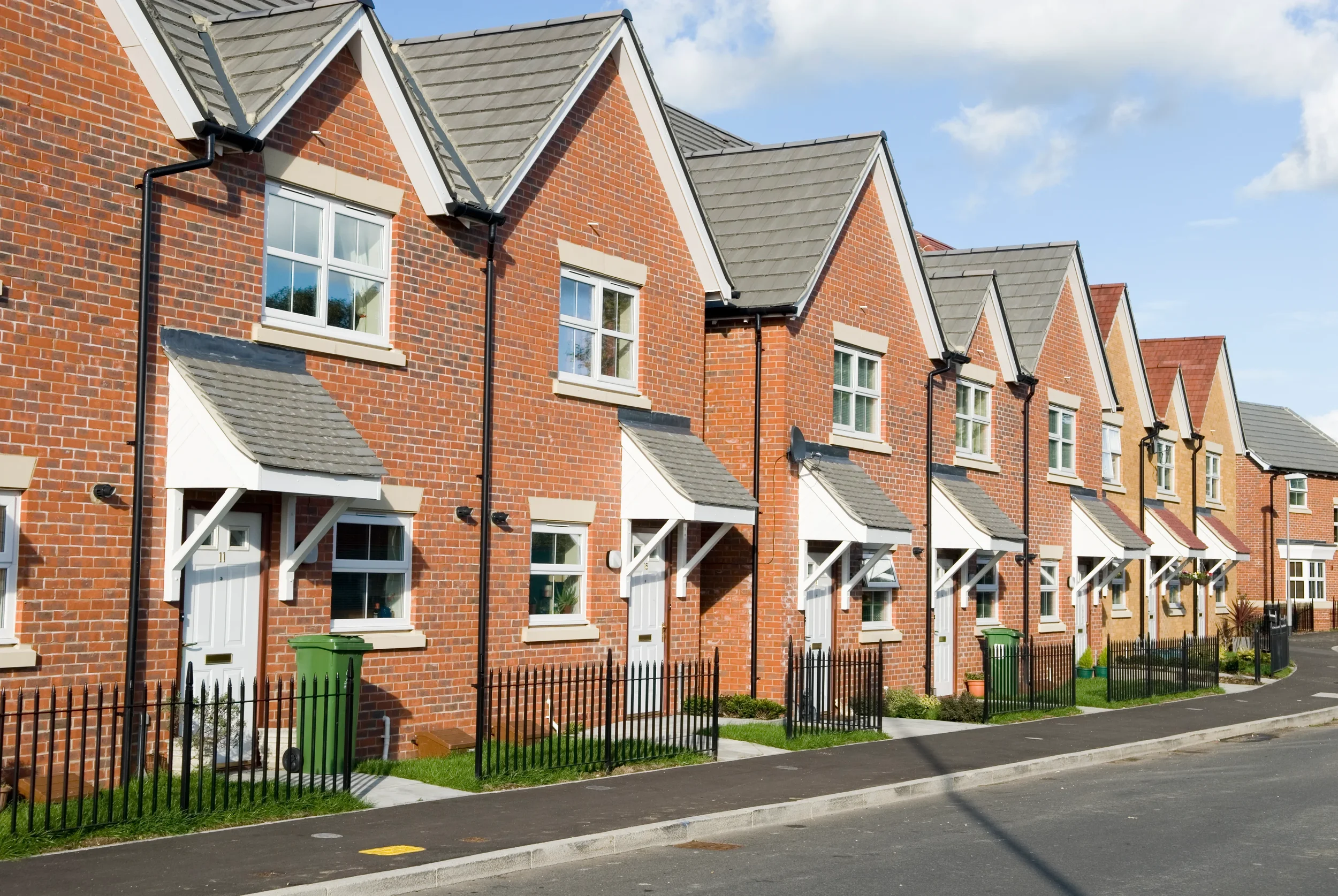 A row of modern brick townhouses with white window frames and small front yards, separated by low black fences, on a paved street under a partly cloudy sky.