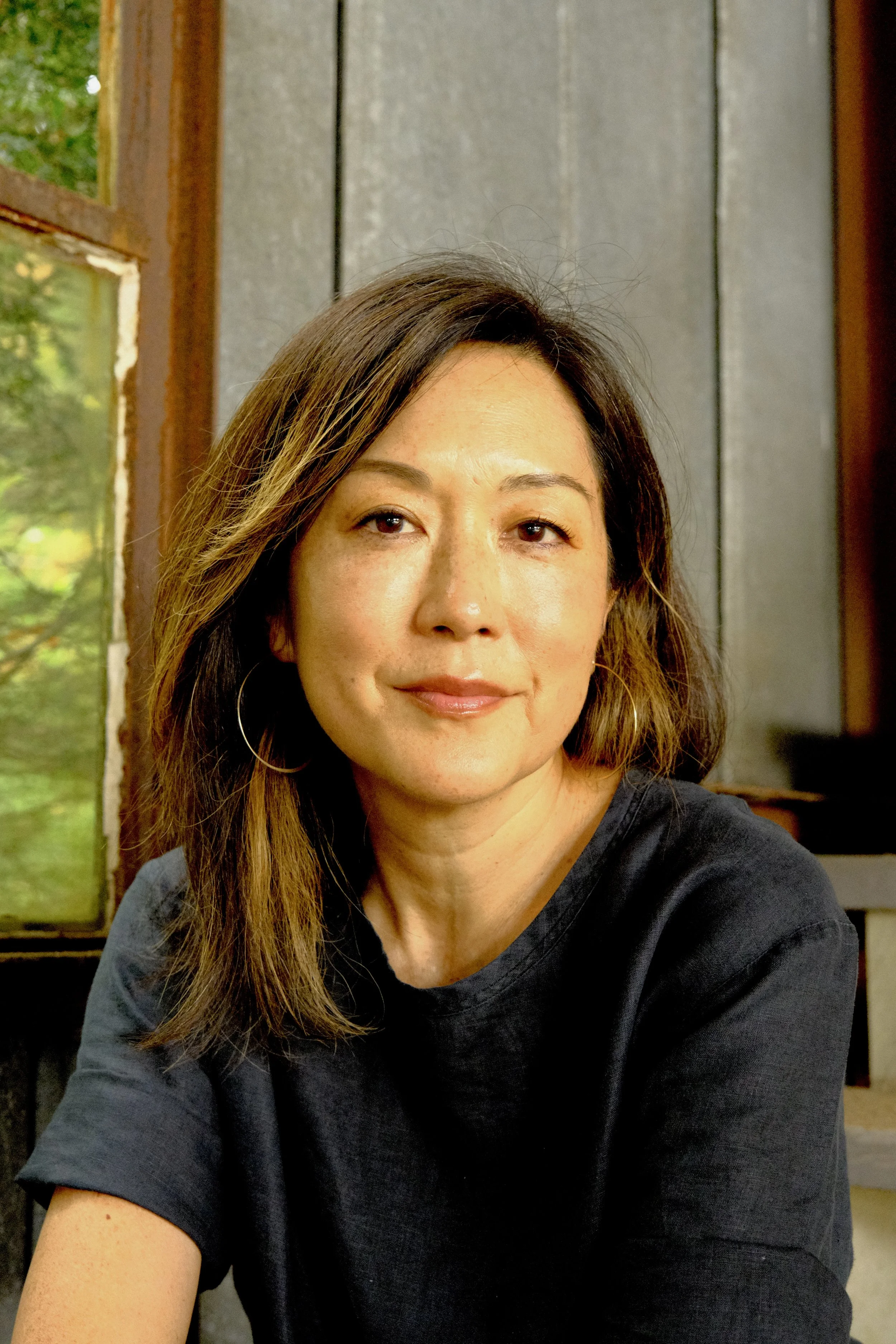 A woman with shoulder-length brown hair, wearing hoop earrings and a black shirt, sitting inside with a window showing green trees in the background.