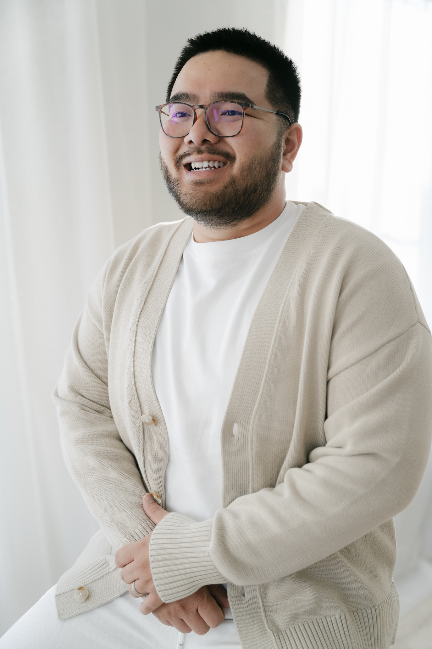 Person smiling, wearing glasses and a beige cardigan, seated in a bright room.