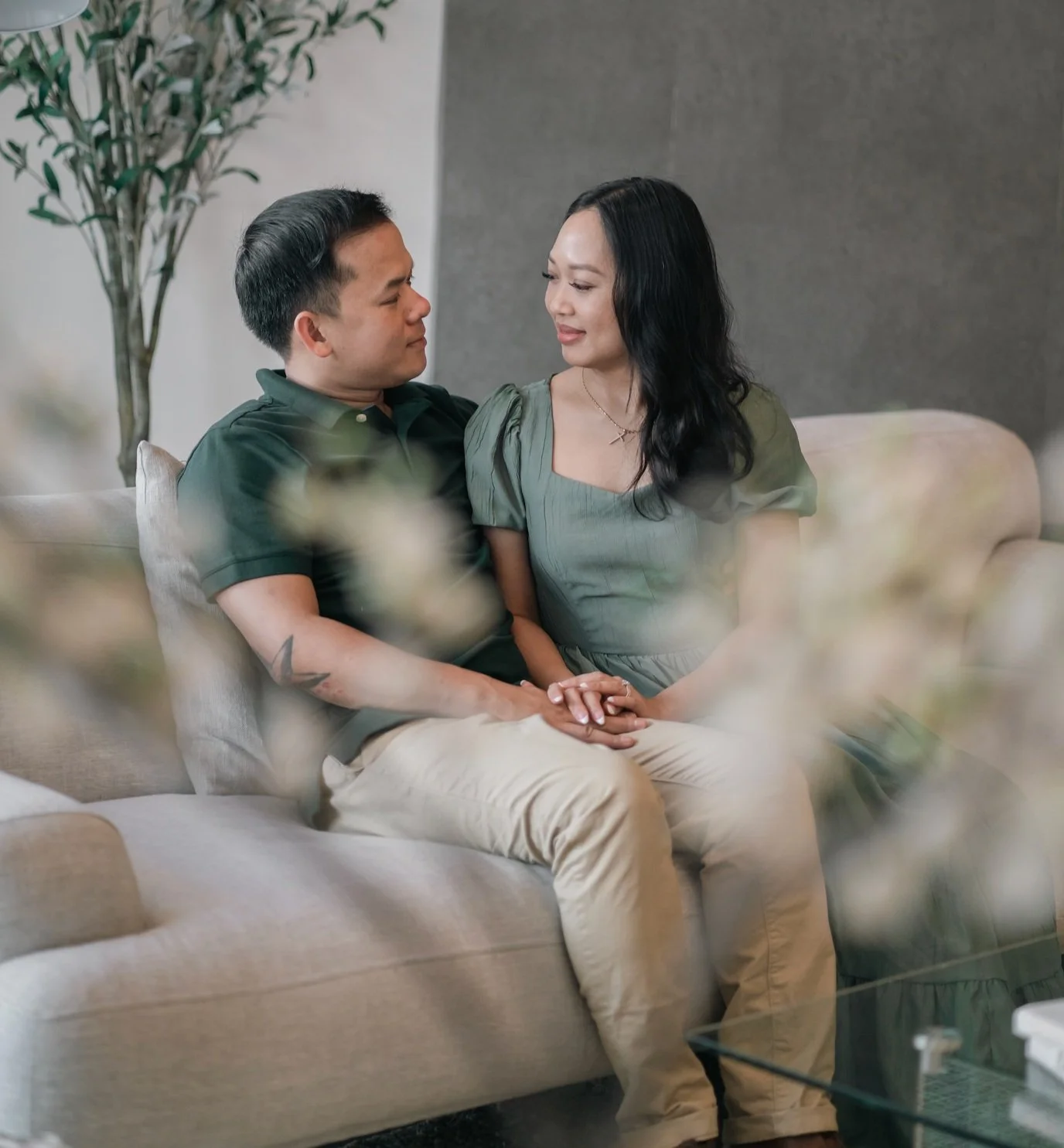 Couple sitting on a couch holding hands and looking at each other, with decorative plants in the background.