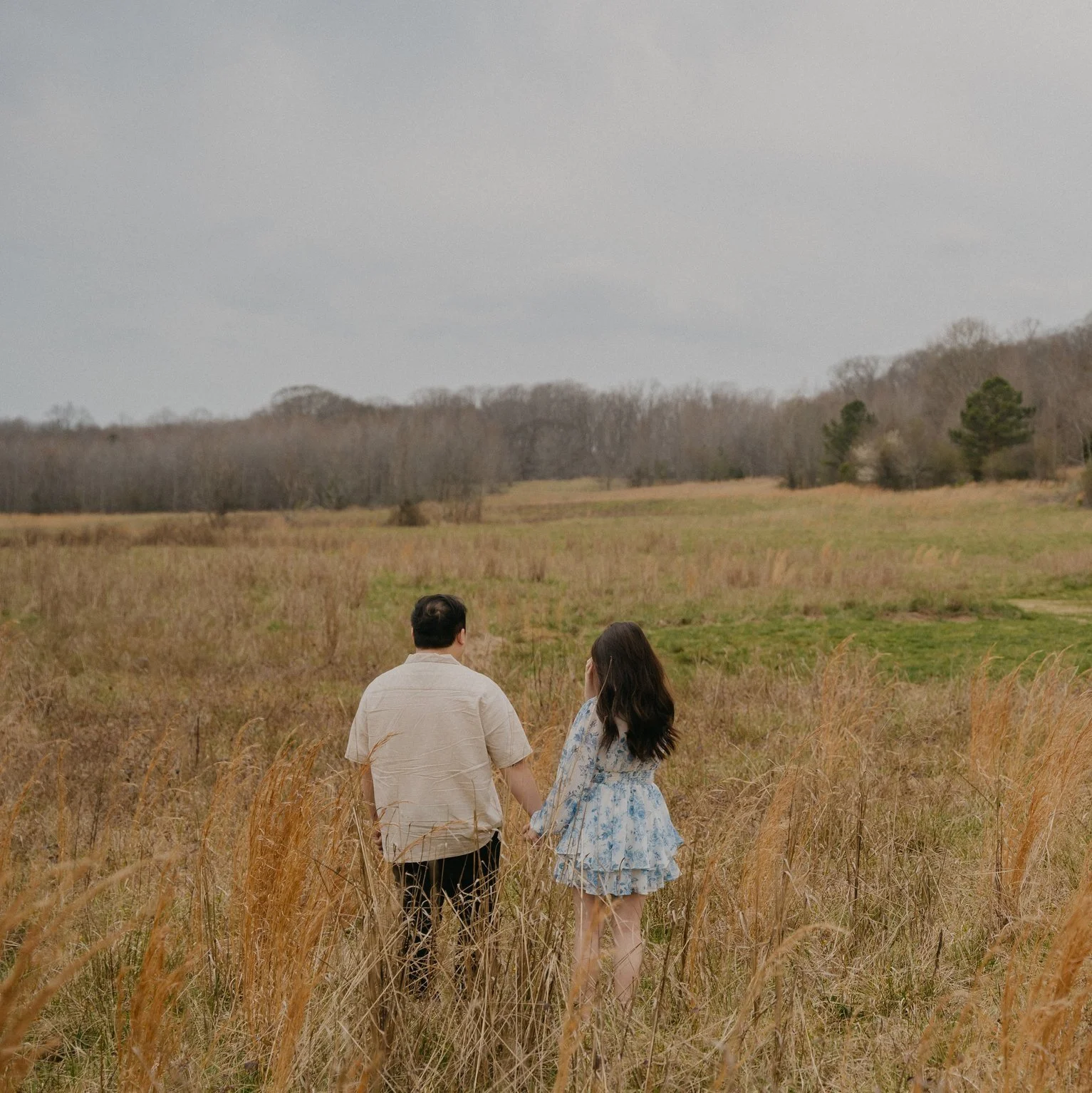 A couple holding hands and walking through a grassy field with trees in the background.