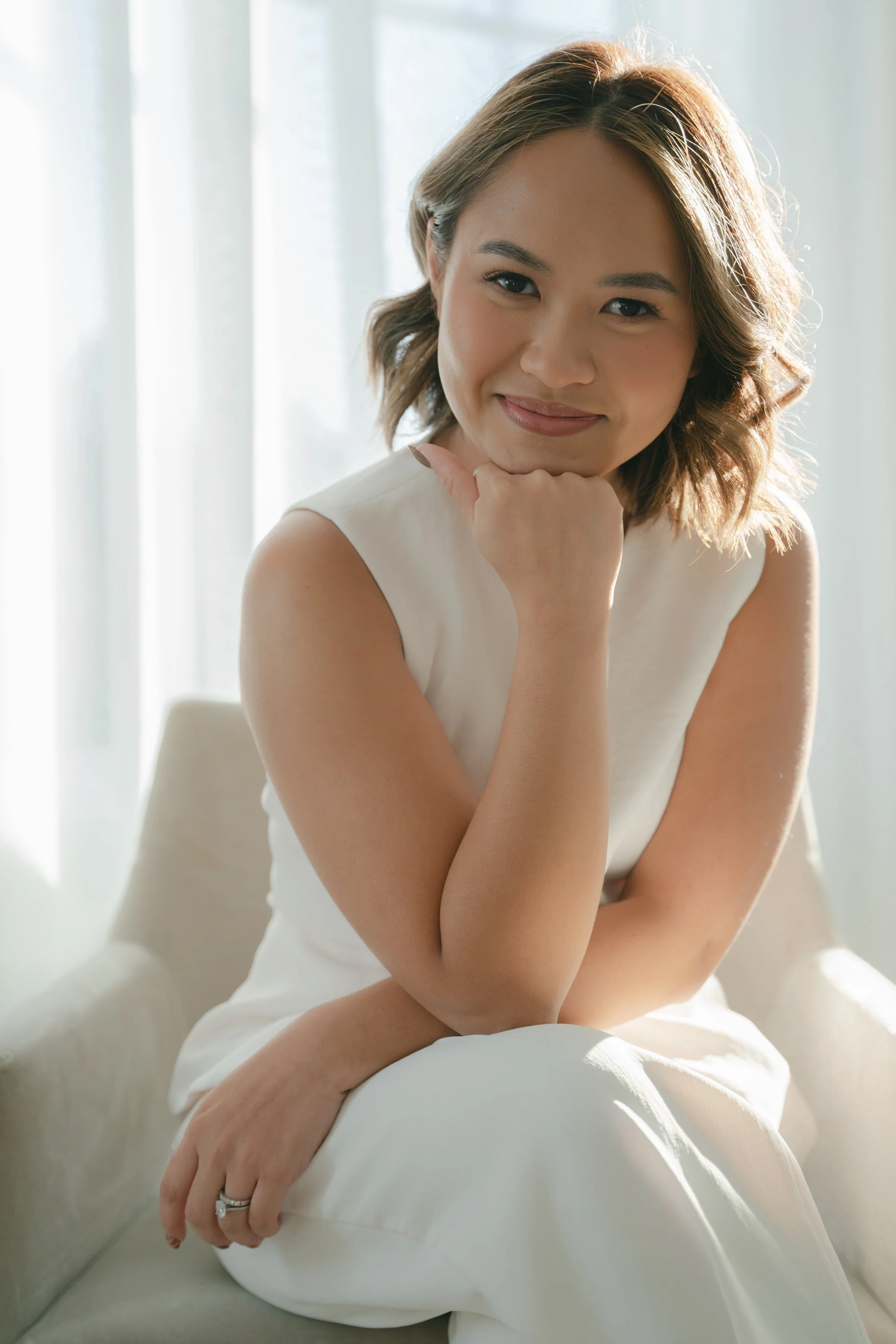 Woman sitting on a chair, wearing a white sleeveless outfit, looking at the camera with a hand resting on her chin, in a softly lit room.