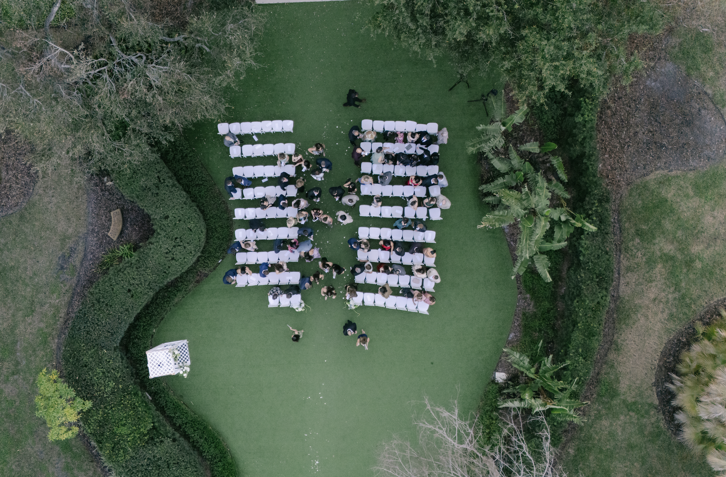 Aerial view of an outdoor wedding ceremony with white chairs and guests seated on a green lawn surrounded by trees and shrubs.