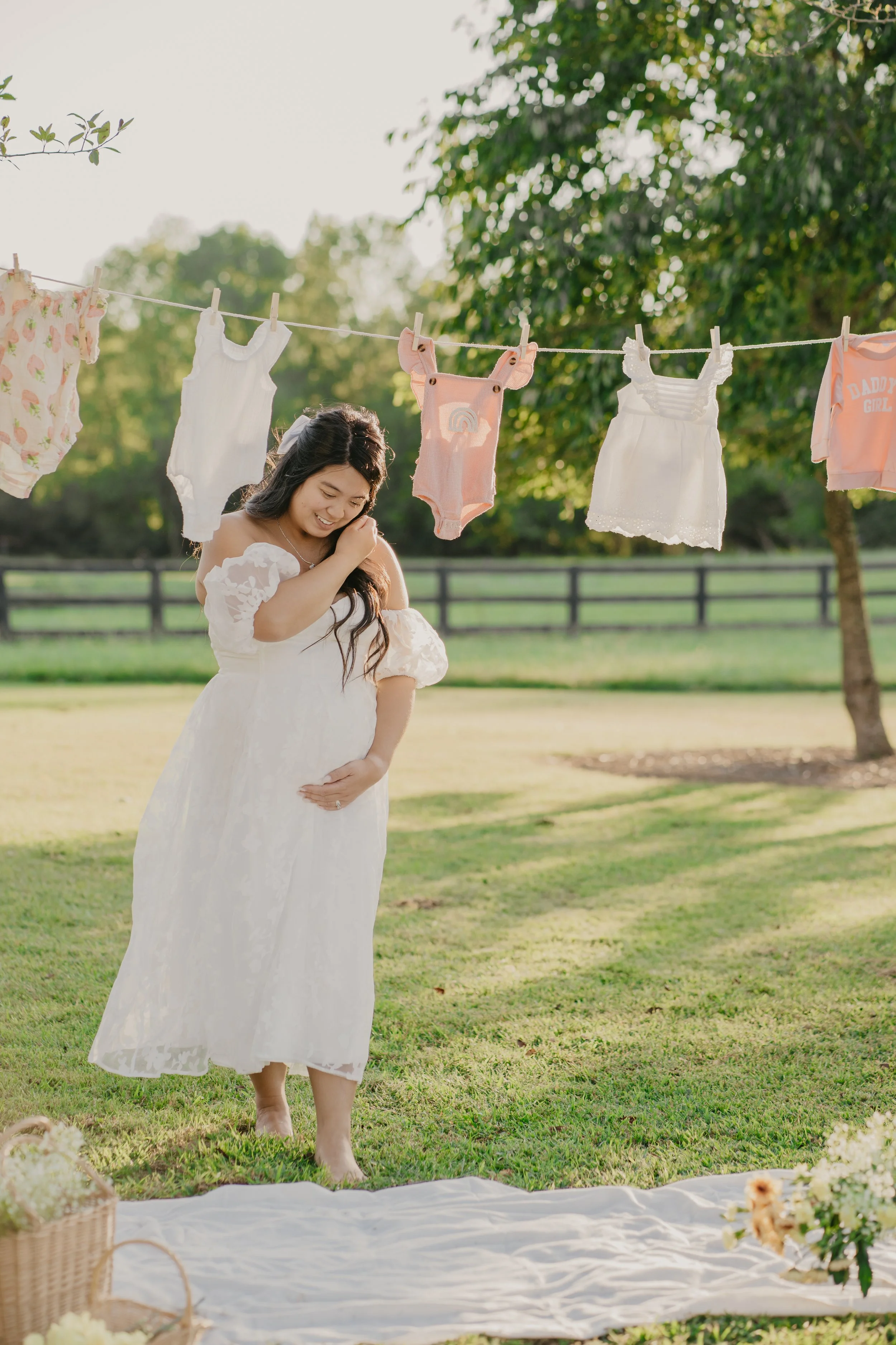 Pregnant woman in white dress touching her belly, standing outdoors under a clothesline with baby clothes, in a grassy garden area.