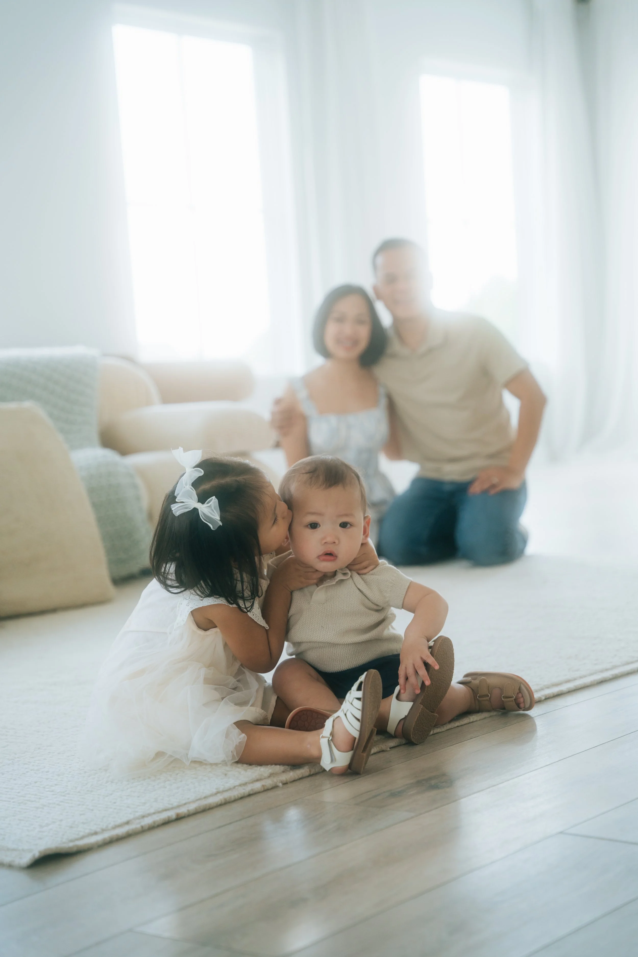 A young girl kisses a baby on the head while sitting on a rug in a living room with parents seated in the blurred background.