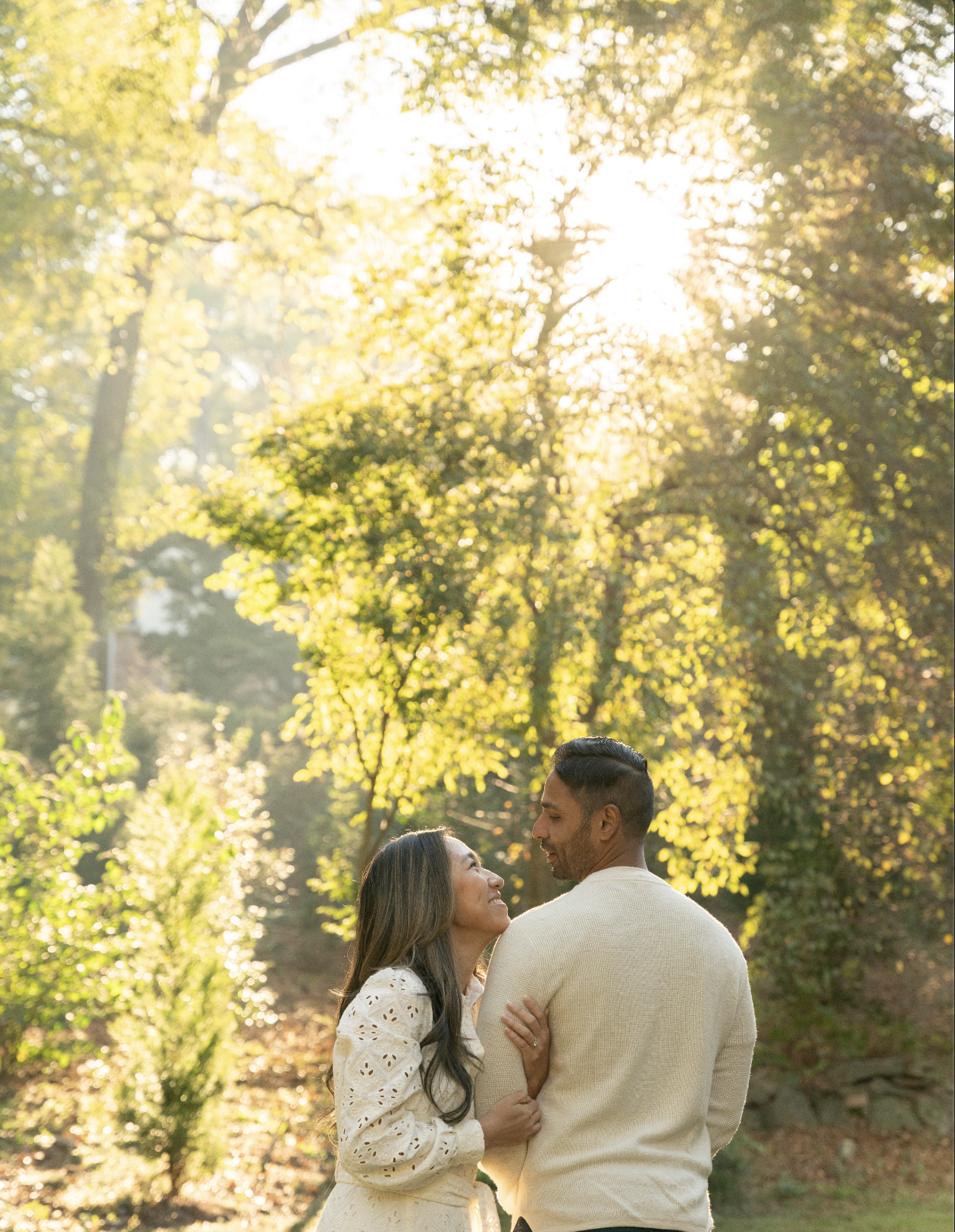 A couple embracing in a sunlit forest with lush greenery and trees in the background.