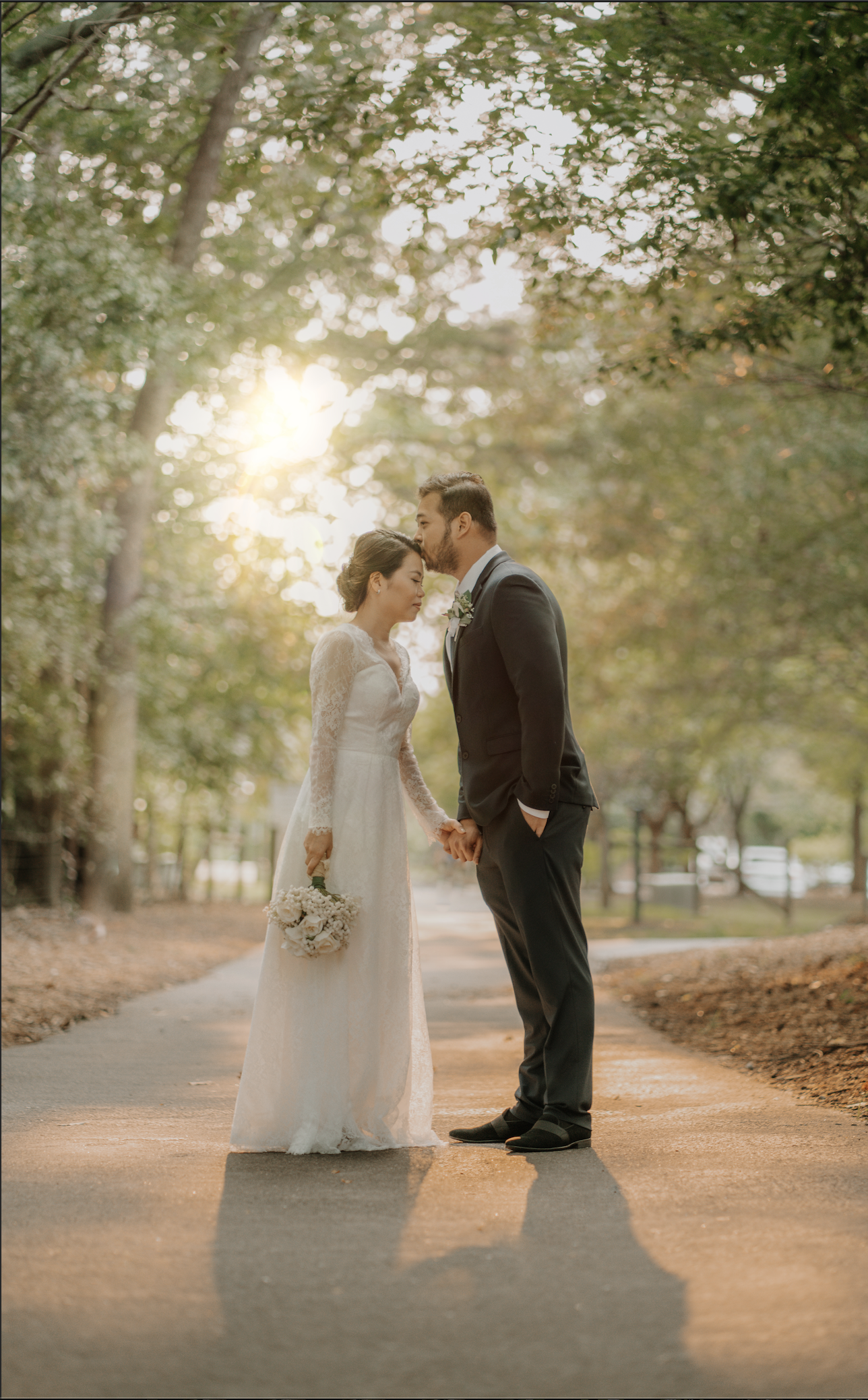 Bride and groom holding hands in a sunlit wooded path, bride wearing a white gown and holding flowers, groom in a suit, leaning down to kiss her forehead.