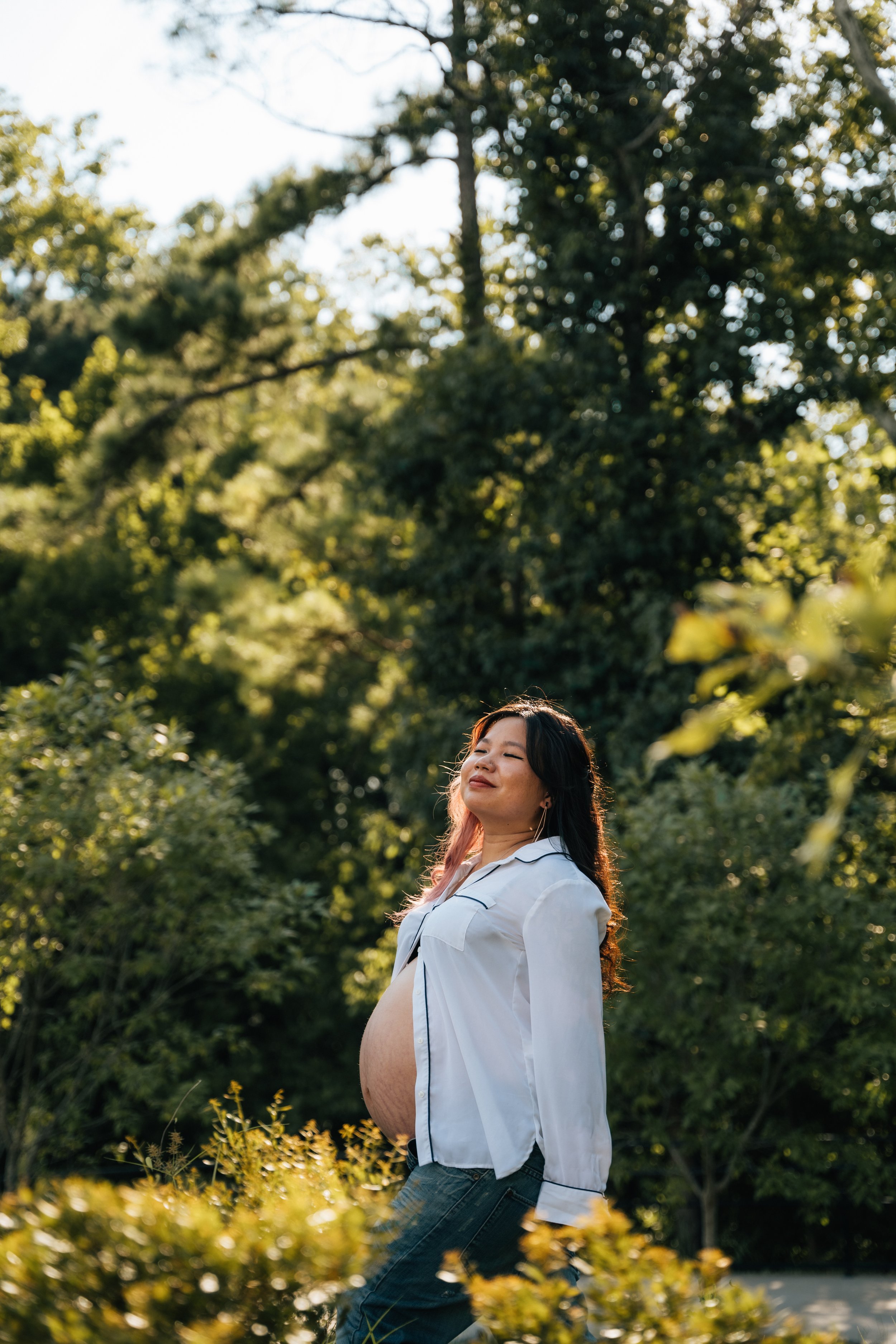 Pregnant woman in a white shirt standing in a sunlit forest