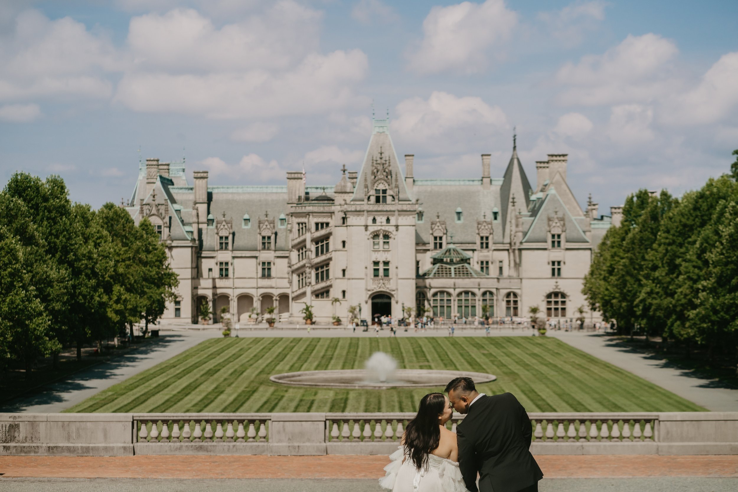 A couple in formal attire stands in front of a large historic mansion with landscaped gardens.