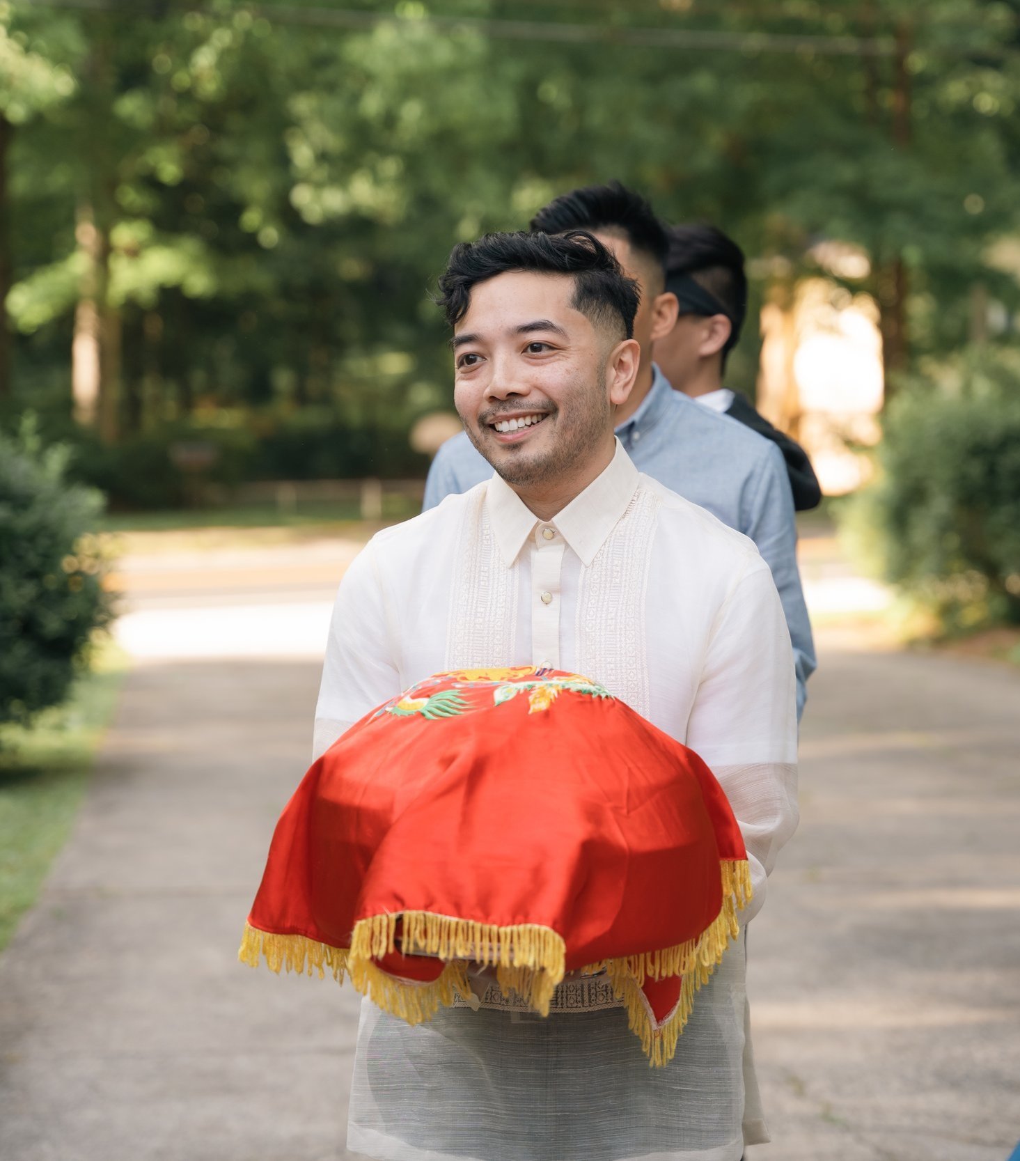 A smiling person dressed in traditional attire holding a red cloth-covered item, outdoors with trees in the background.