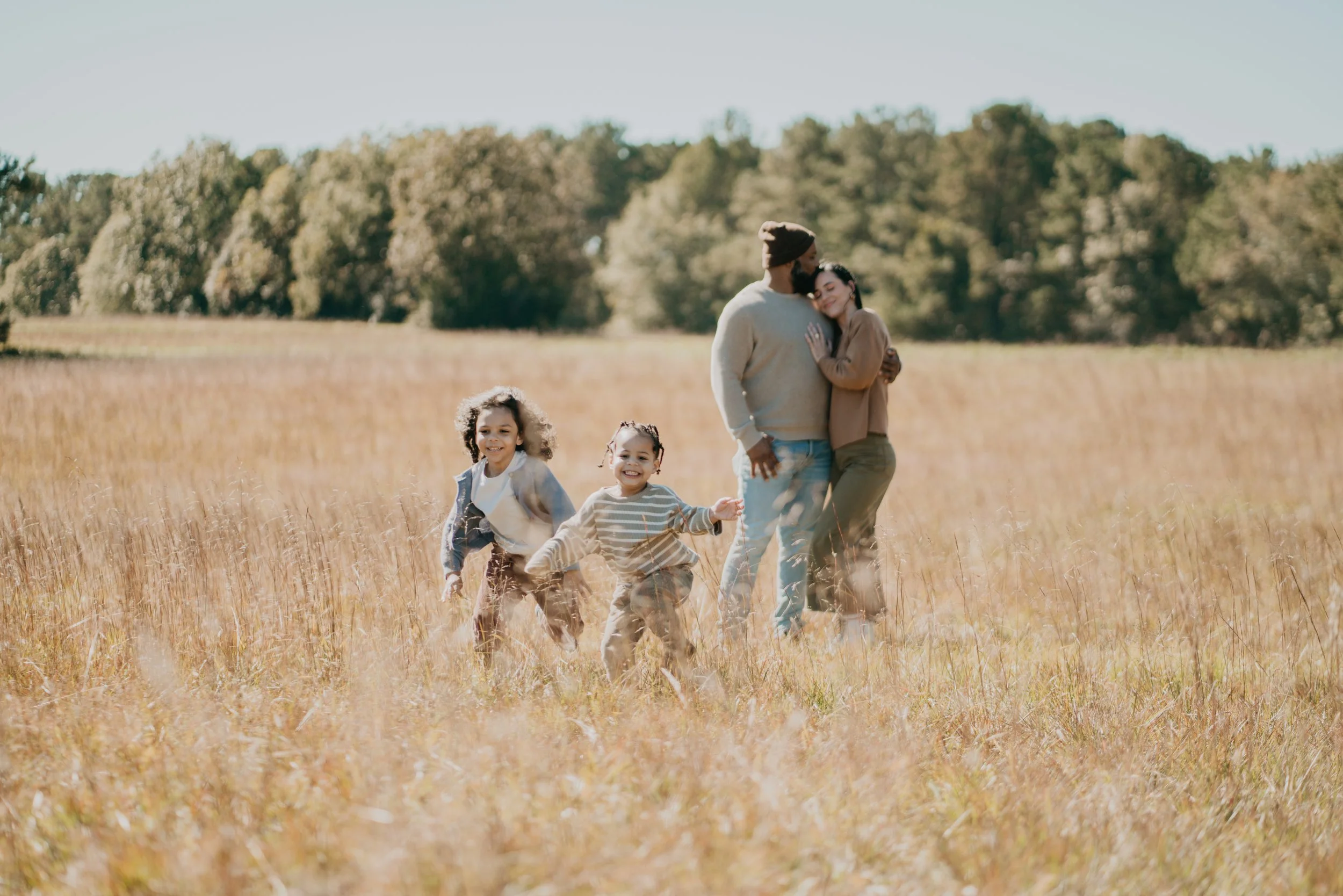 Family playing in a grassy field with trees in the background; two children running in the foreground and parents hugging in the background.