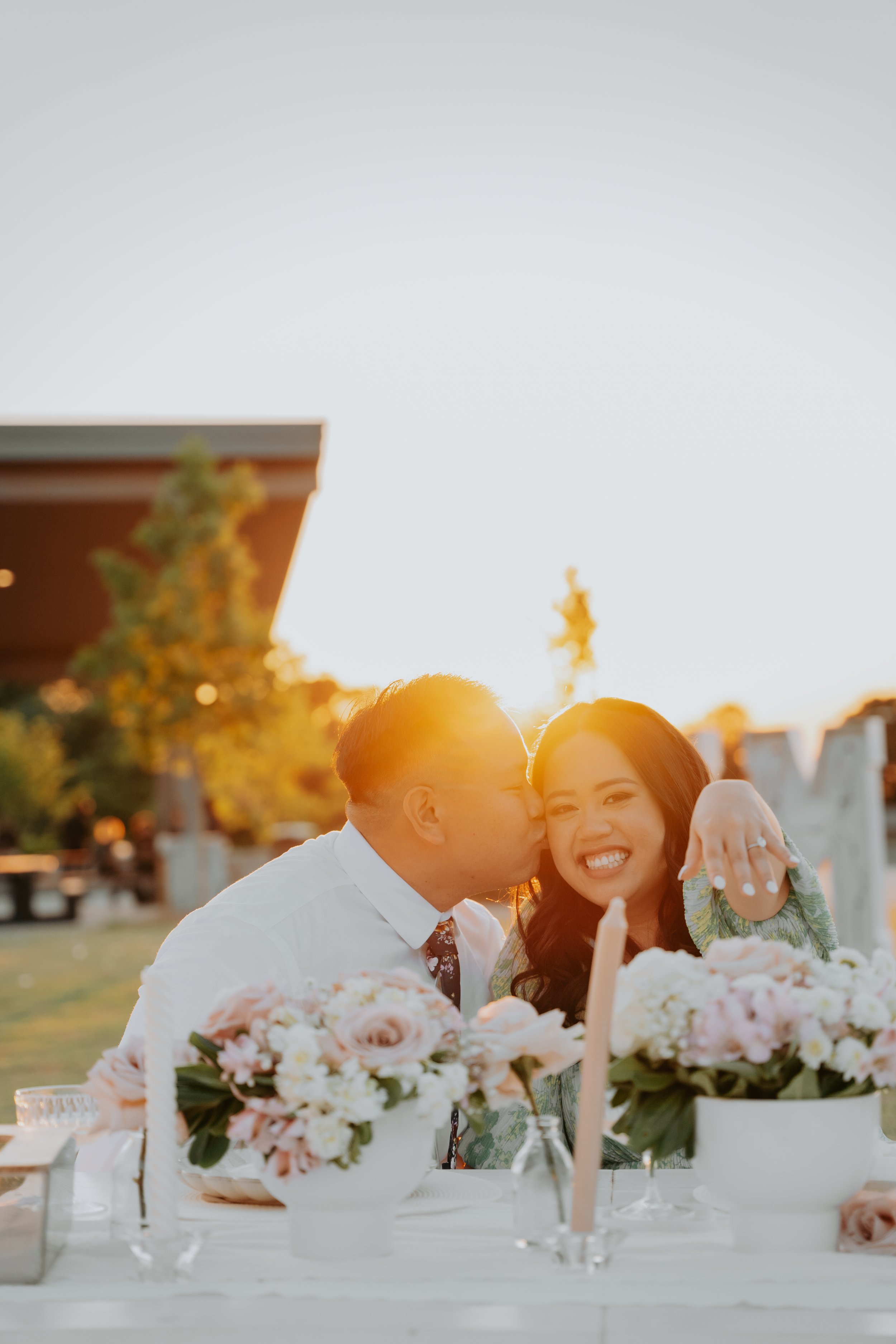 A couple sitting at a table decorated with flowers, with the man kissing the woman on the cheek. She is smiling and showing an engagement ring on her hand. The sun is setting in the background.
