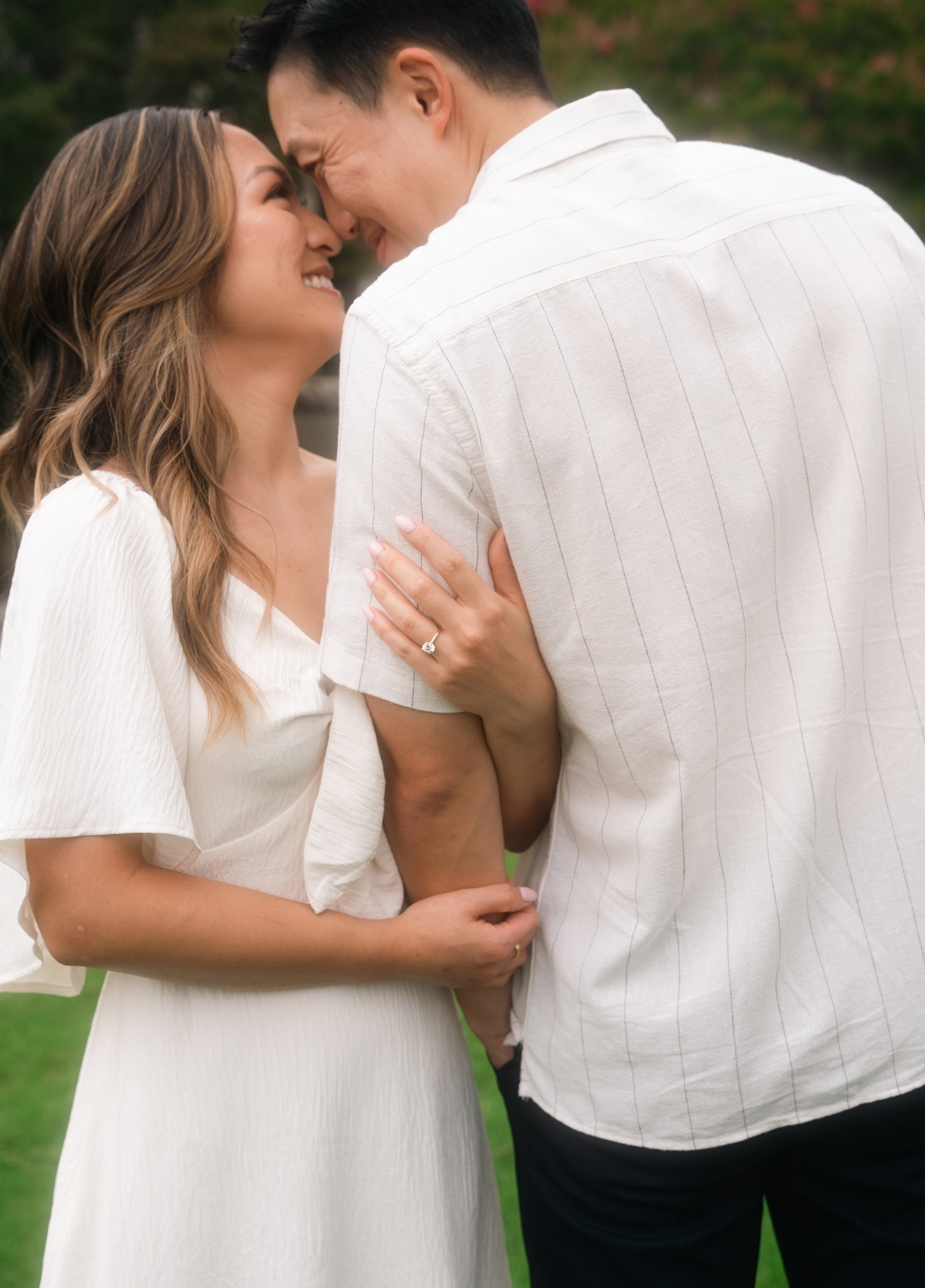 Happy couple embracing, wearing white outfits, with woman showing an engagement ring.