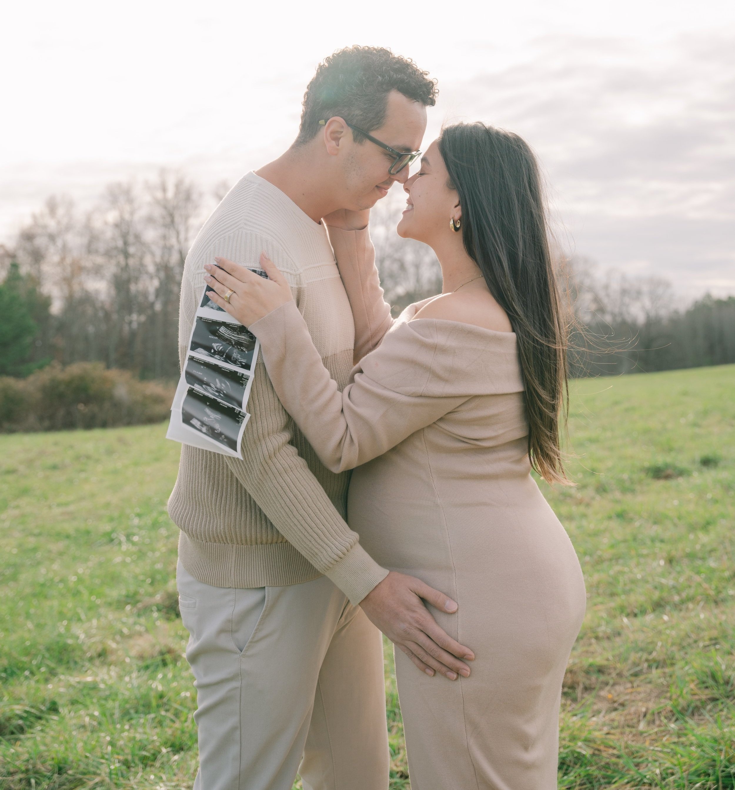 A pregnant couple embracing outdoors, holding ultrasound photos.