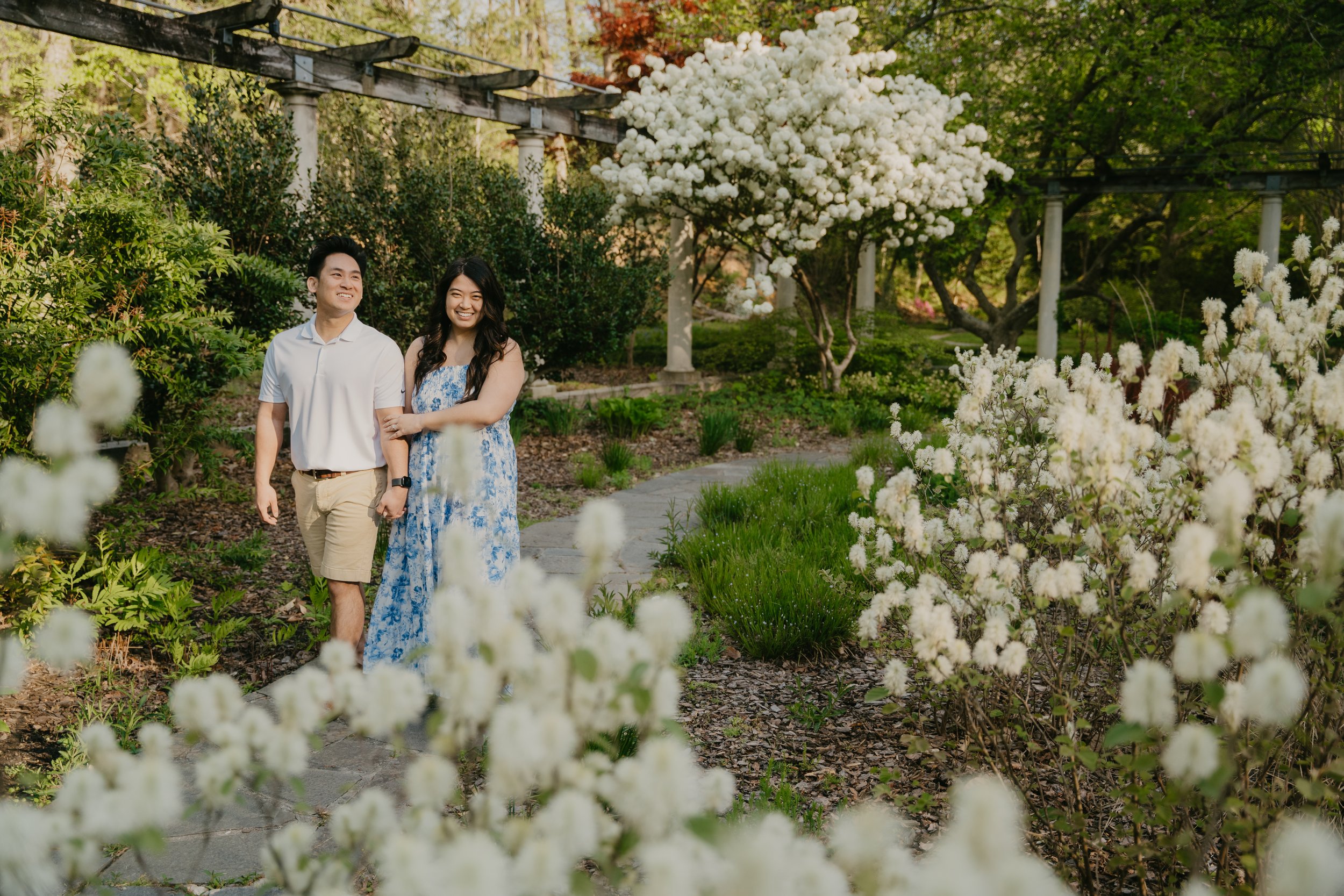 Couple walking through a garden pathway surrounded by blooming white flowers and green foliage.