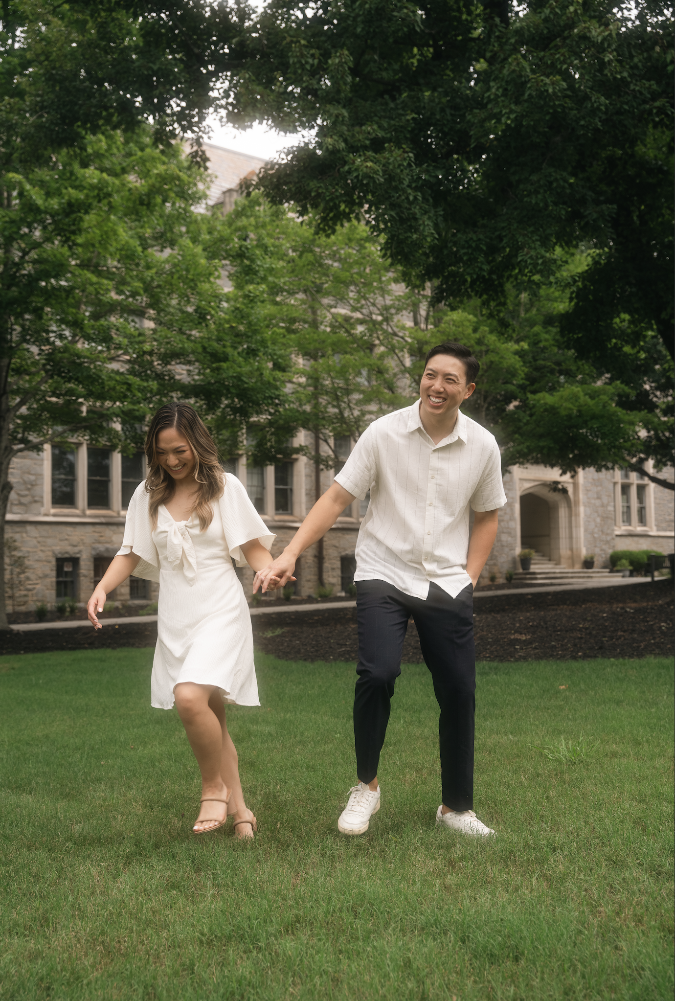 A man and woman holding hands and walking on grass, near a stone building, surrounded by lush green trees.