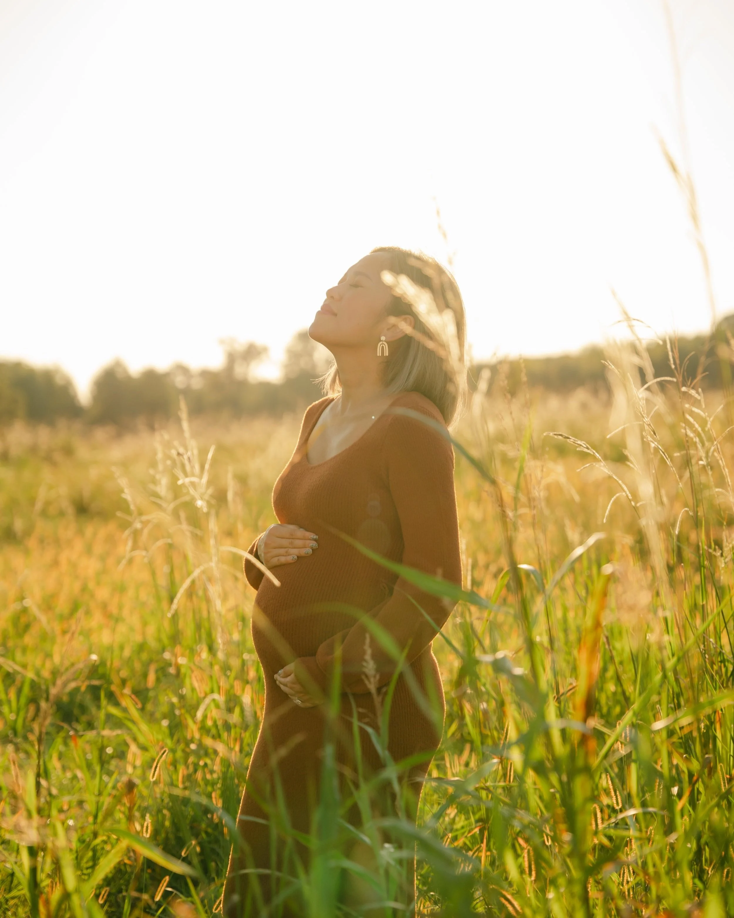 Pregnant woman standing in a sunlit field, holding her belly, with eyes closed and smiling.