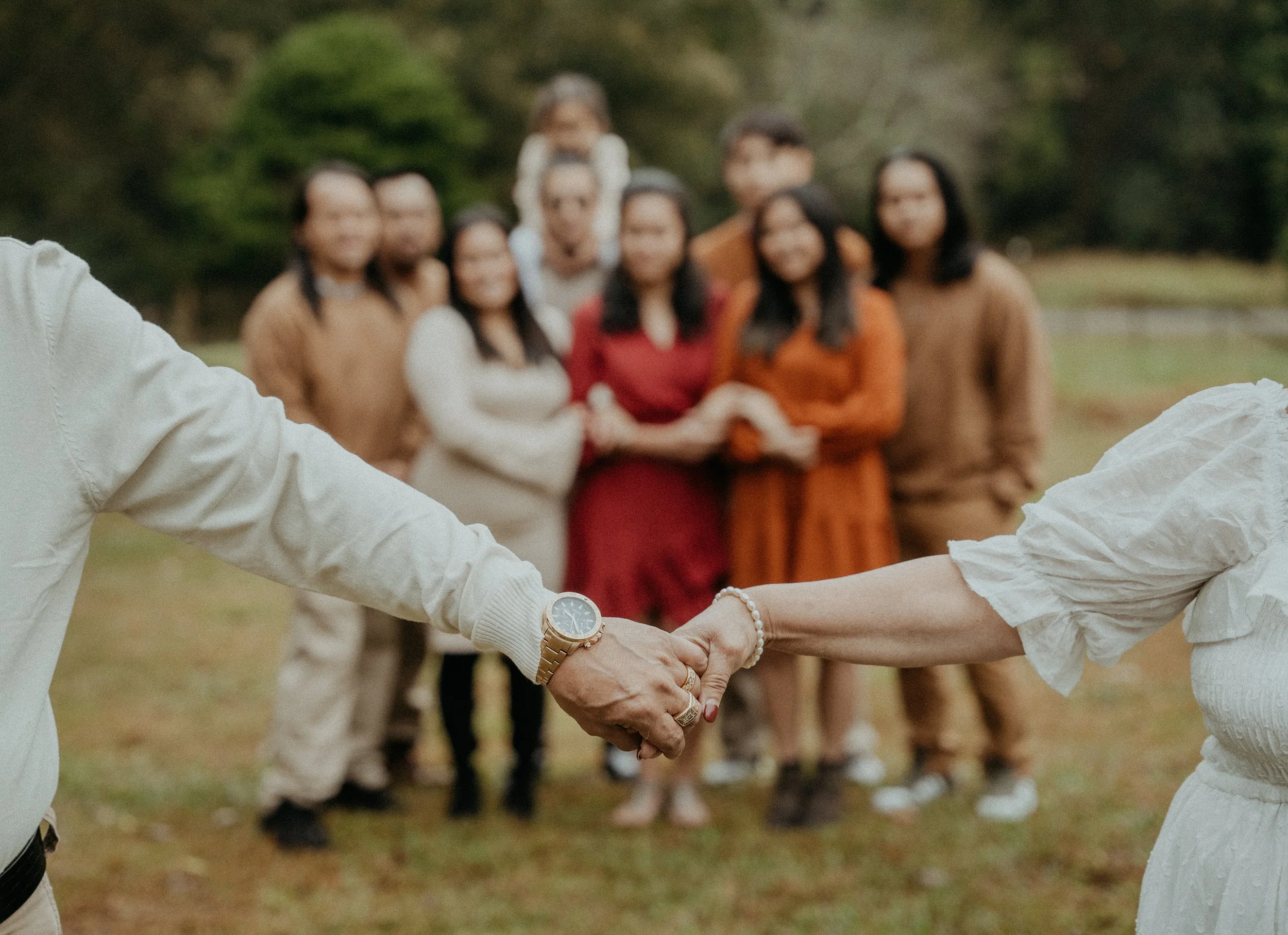 A man and woman holding hands in the foreground wearing white clothing and jewelry, with a group of people in the blurred background standing in a park.