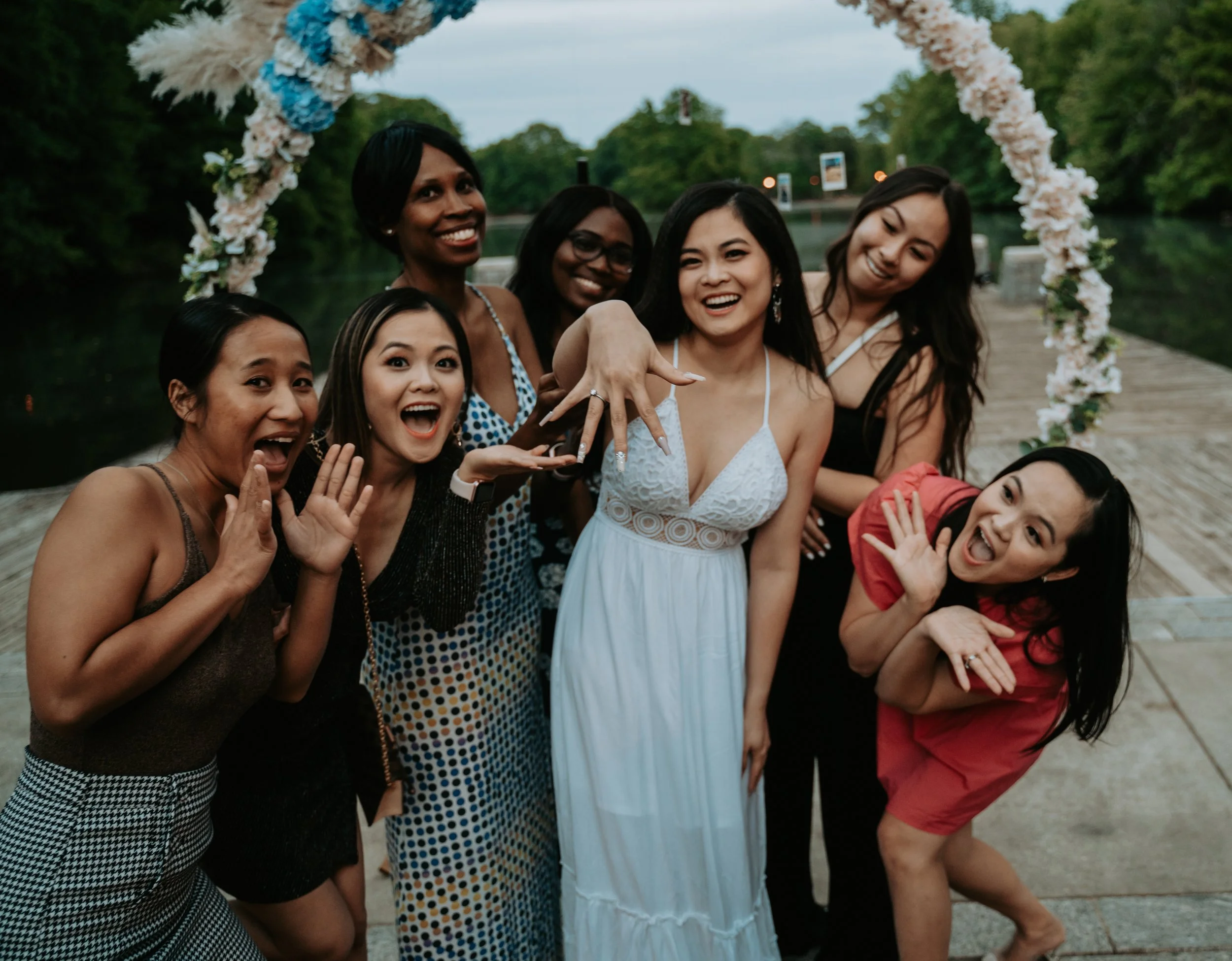 A group of women celebrating an engagement, with one prominently showing an engagement ring. They are outdoors under a floral arch near a body of water, and everyone is smiling and expressing excitement.