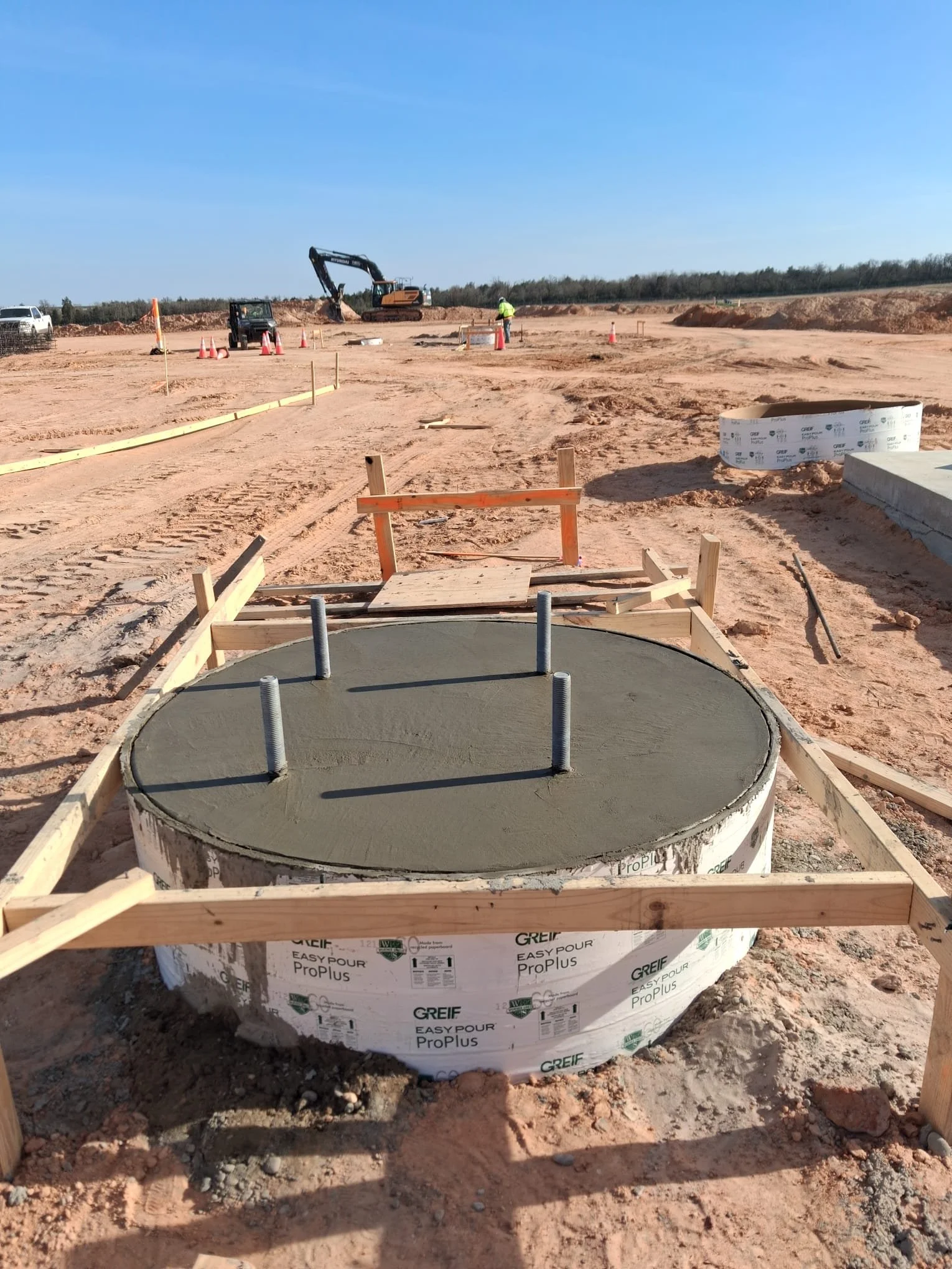 Construction site with a circular concrete foundation with metal bolts, wooden framing, and construction workers and machinery visible in the background.