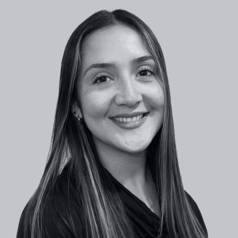 Black and white portrait of a young woman with long straight hair, smiling with visible teeth, wearing a dark top, against a plain light background.