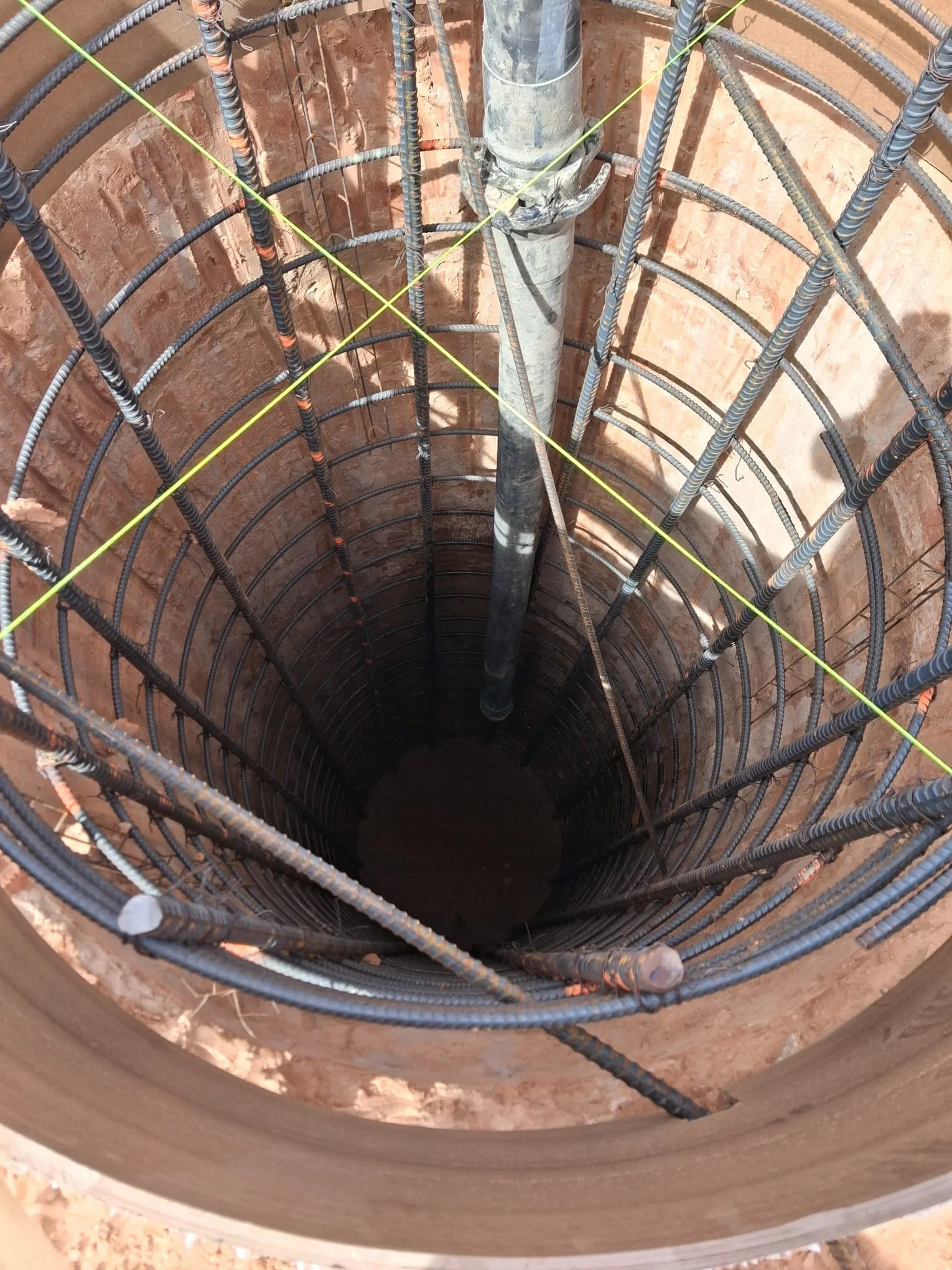 A vertical construction shaft with exposed rebar and a central pipe, lined with brick and wrapped with yellow safety lines.