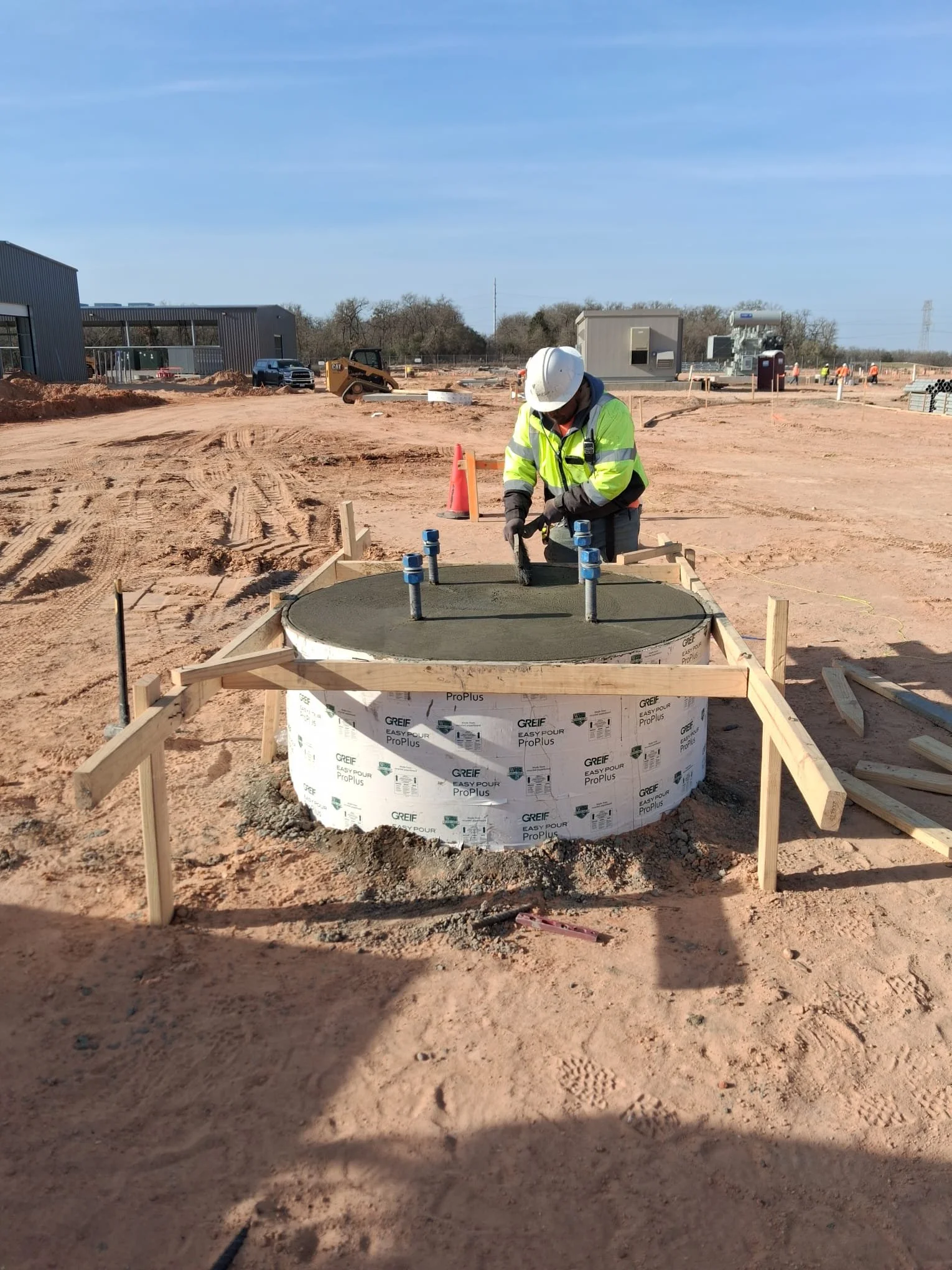Construction worker in a yellow safety vest and white helmet smoothing wet concrete in a large circular mold with blue spacers at a construction site with dirt and construction equipment in the background.