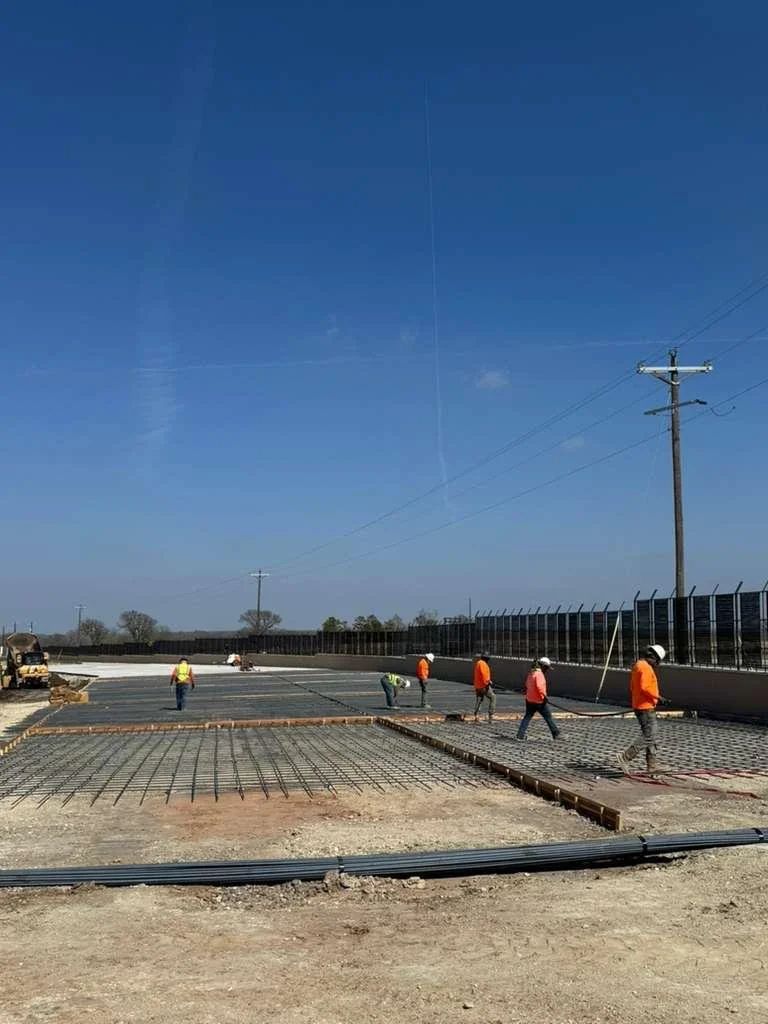 Construction workers in blue helmets spreading wet concrete on a building site with rebar structures and a small excavator in the background.