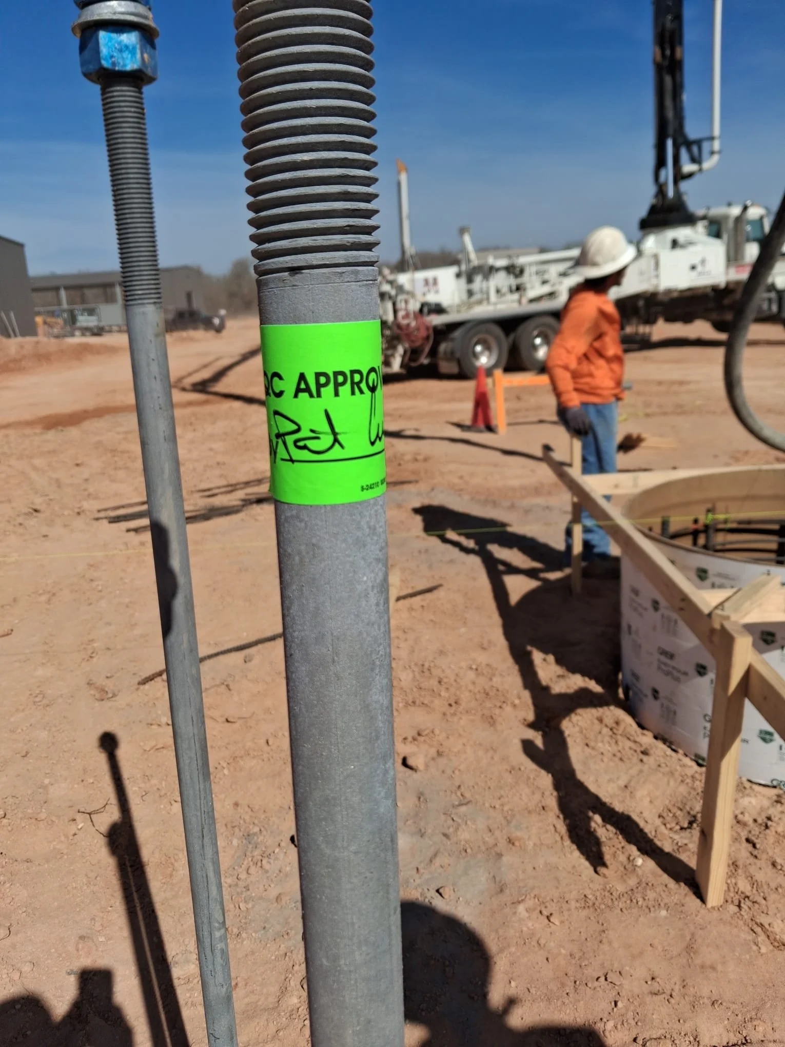 Close-up of a construction site with metal pipes, one with a neon green 'PLC APPROVAL' sticker, and a worker wearing a white helmet and orange jacket in the background.