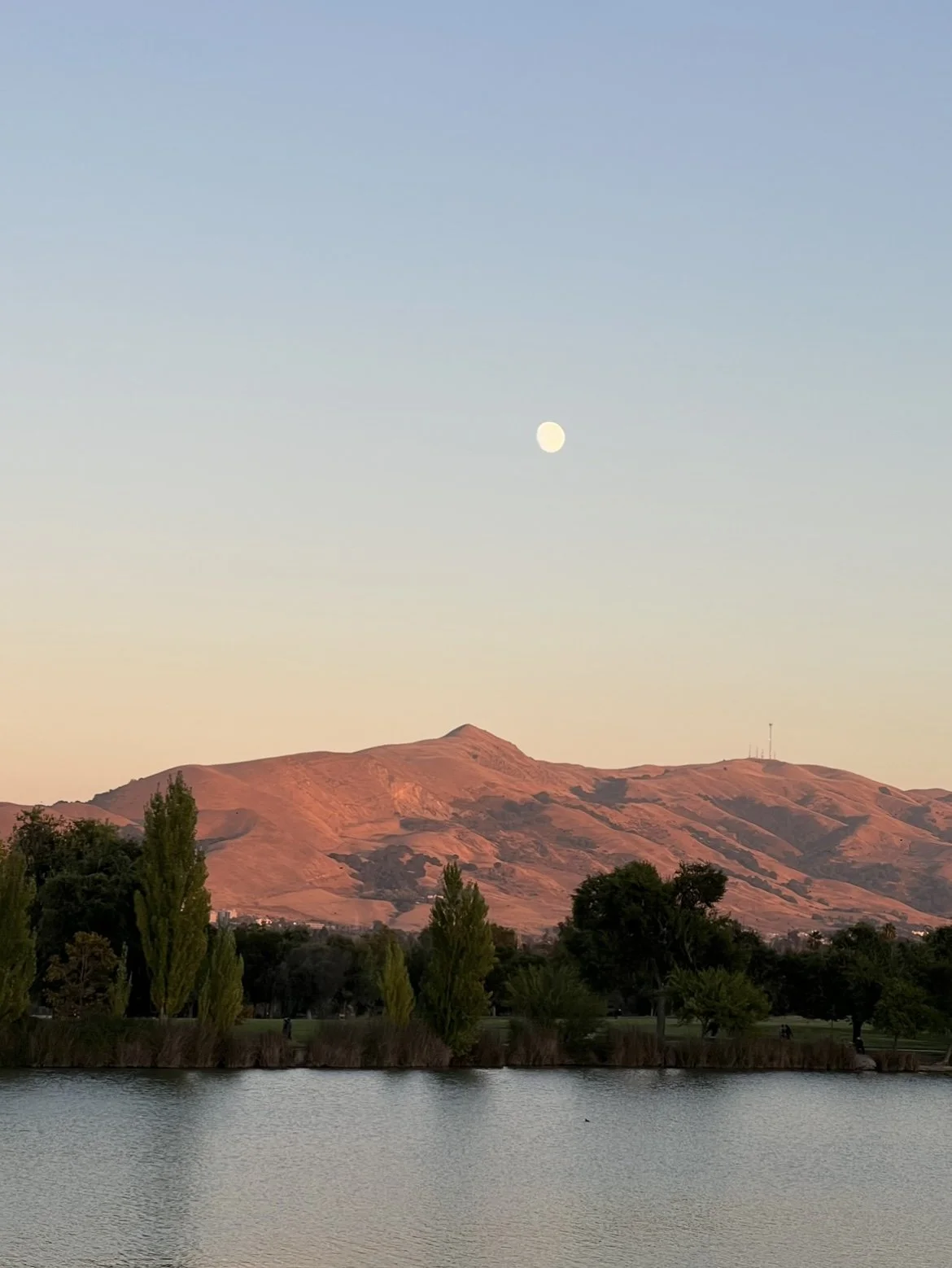 Landscape with a lake, trees, and mountains under a clear sky at sunset with the moon visible.