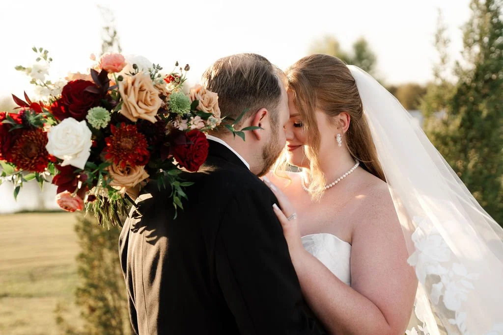 Bride and Groom embrace with bouquet of deep red and gold flowers