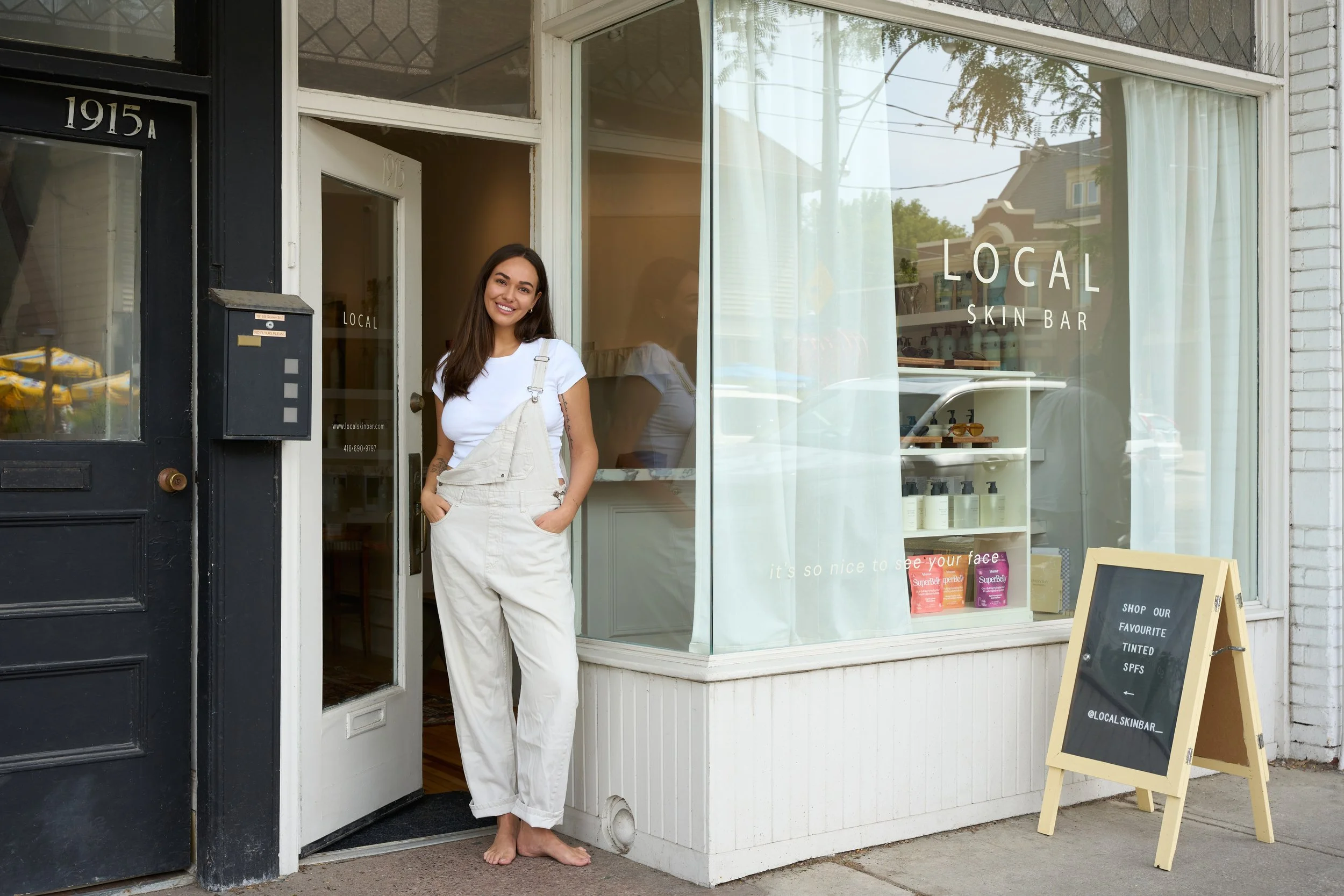 A young woman with long dark hair, wearing a white t-shirt and beige overalls, standing barefoot outside a skin care shop called 'Local Skin Bar,' smiling at the camera.