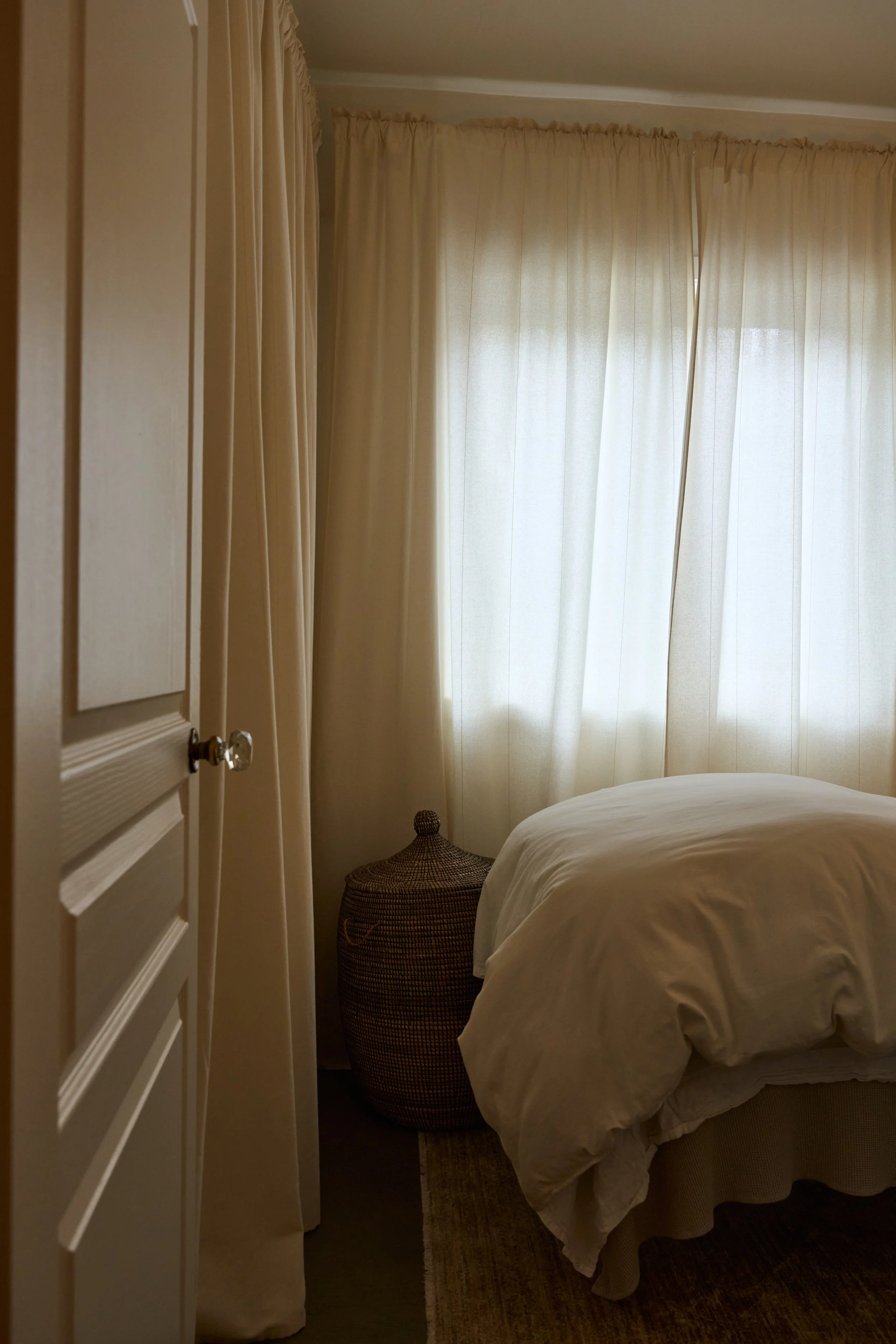 A cozy facials and skincare treatment room with cream-colored curtains covering a window, a bed with white bedding, a woven basket at the corner of the bed, and dim lighting.