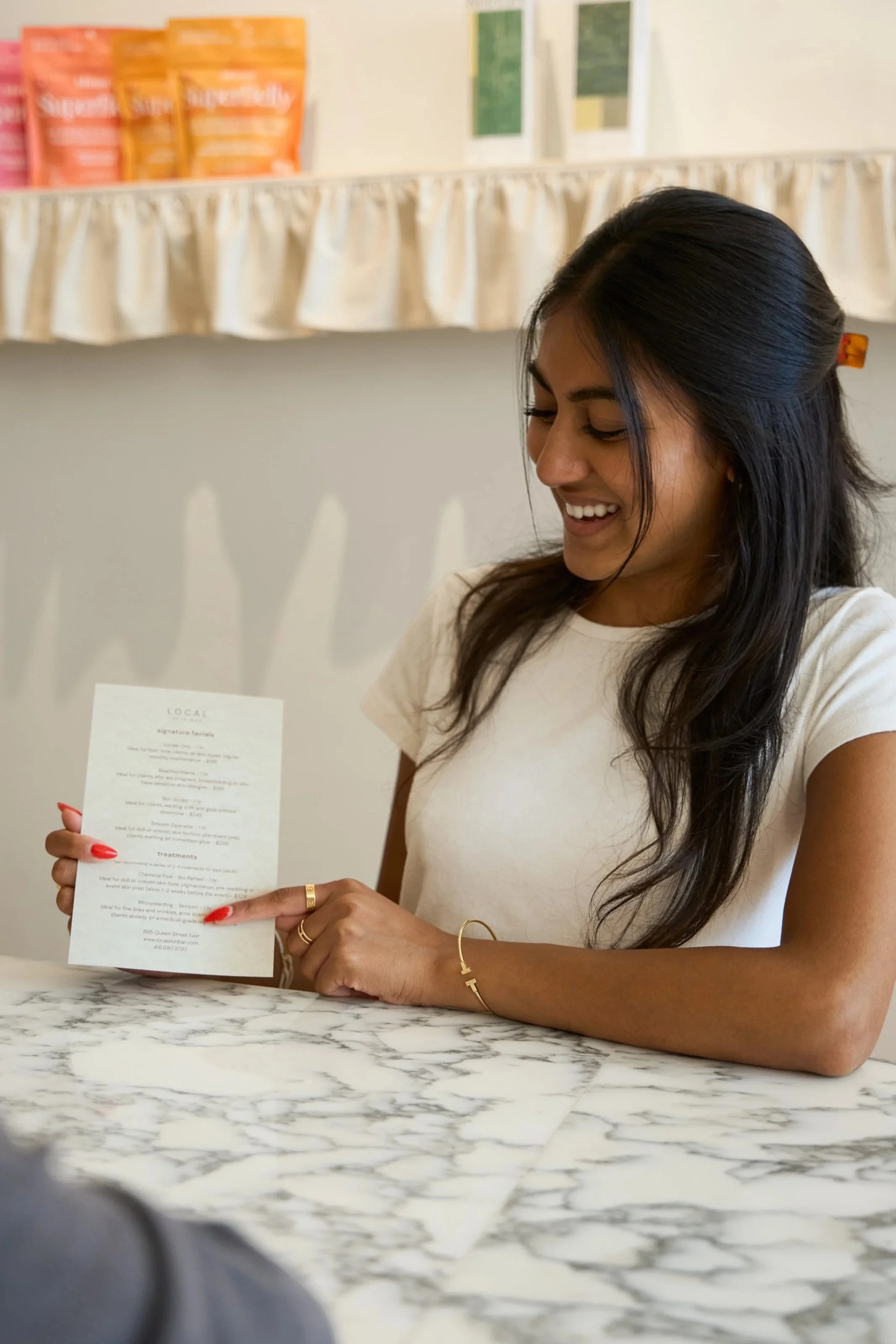 A woman smiling while holding a Local Skin Bar facials menu at a marble counter.