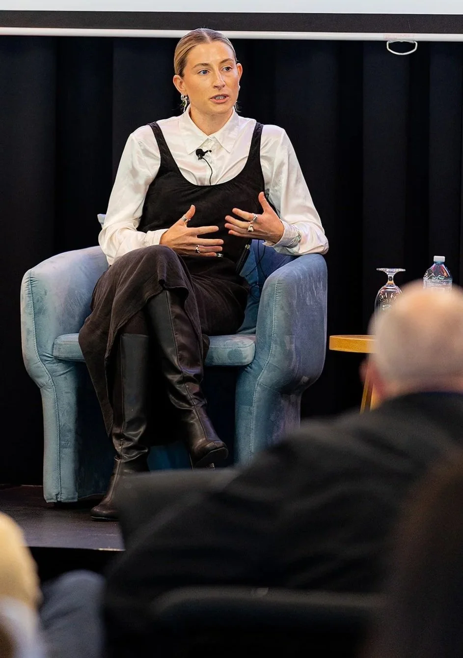 A woman sitting in a light blue armchair speaking during a panel discussion, with audience members visible in the foreground and a black curtain backdrop.