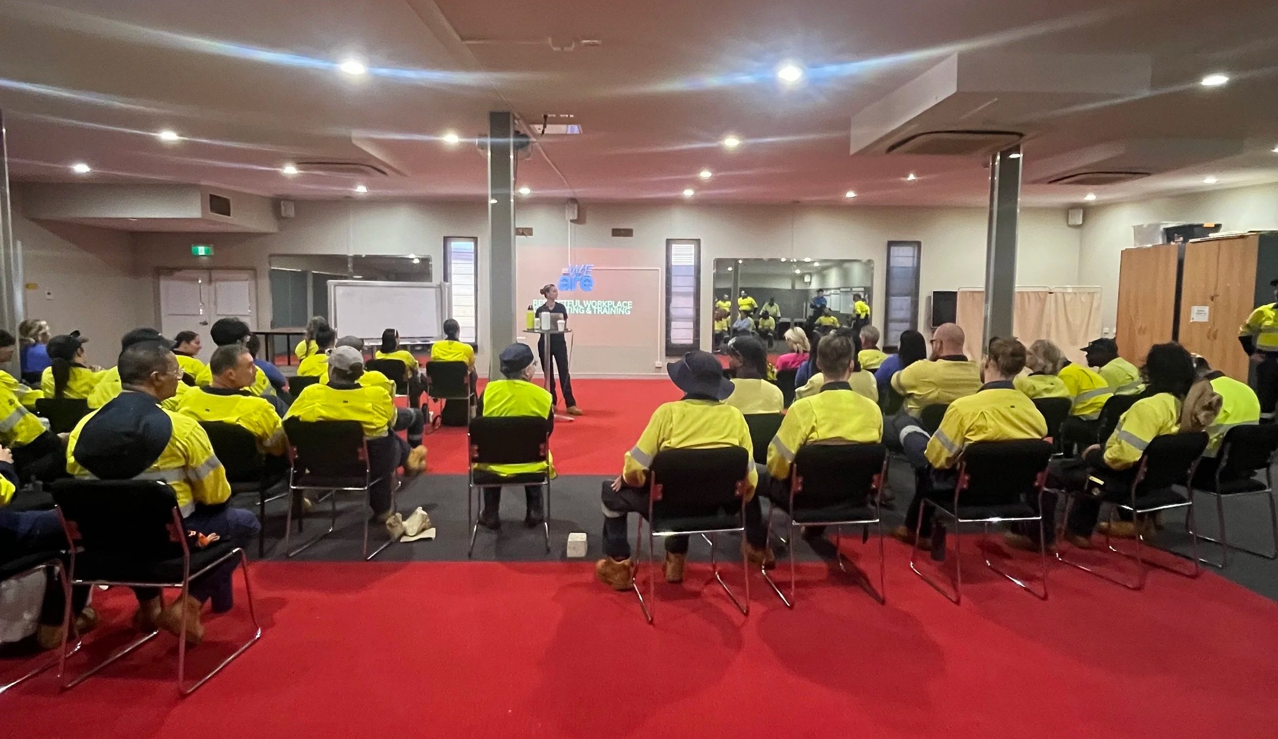 A large group of people in yellow safety vests attending a presentation in a conference room with a red carpet, seated facing a speaker at the front. There are windows, mirrors, and folding partitions along the walls.