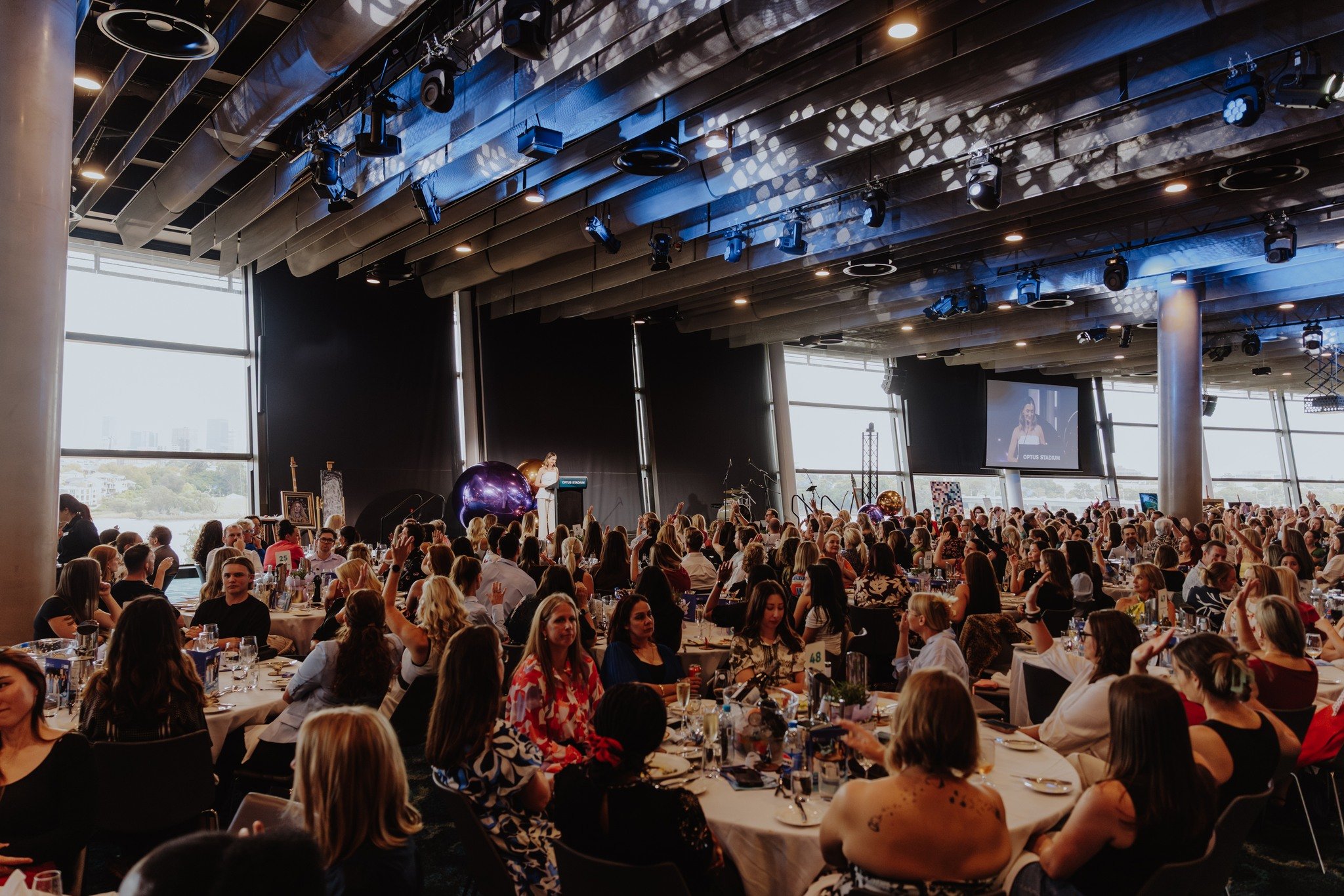 A large indoor event or conference with many attendees seated at round tables, a stage with large screens, and professional lighting, possibly a gala or awards ceremony.