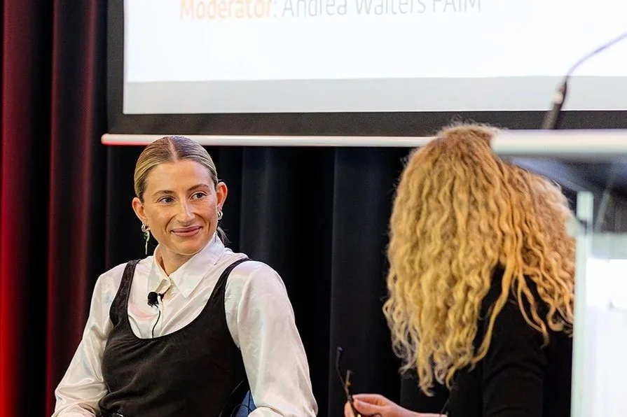 Two women sitting and talking during a panel discussion or interview. One woman has blonde curly hair, facing away, and the other woman has straight blonde hair, smiling, wearing a white shirt and a black dress.