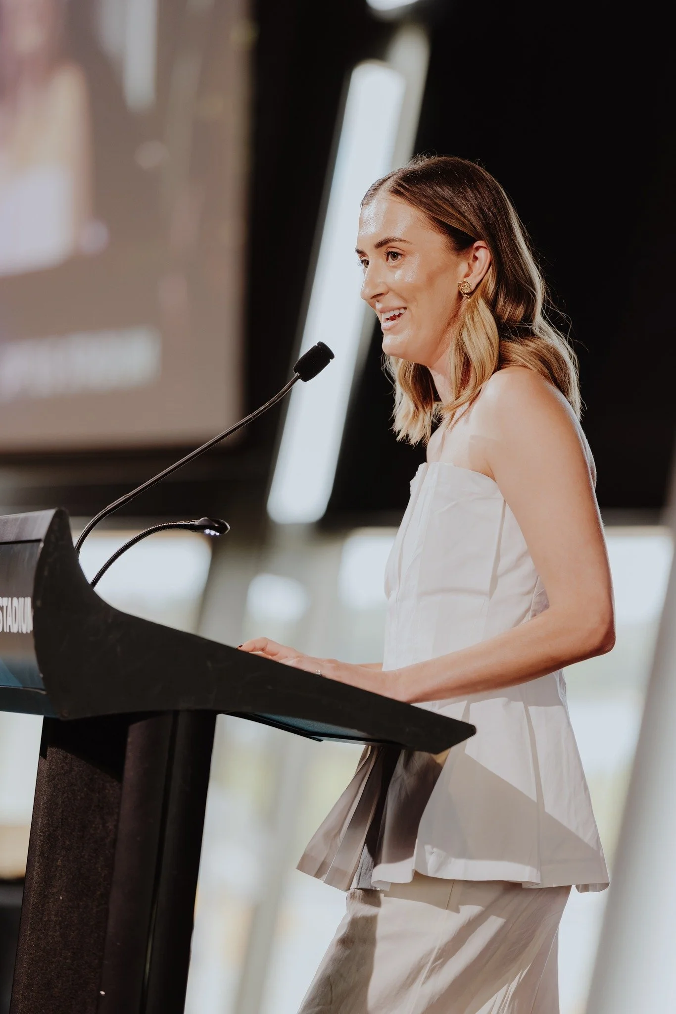 A woman in a white strapless dress is speaking into a microphone while standing at a podium.