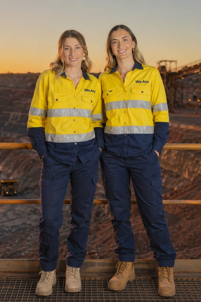 Two women in yellow safety shirts with reflective stripes and navy blue pants standing side by side outdoors at sunset, smiling, with a mining or construction site in the background.