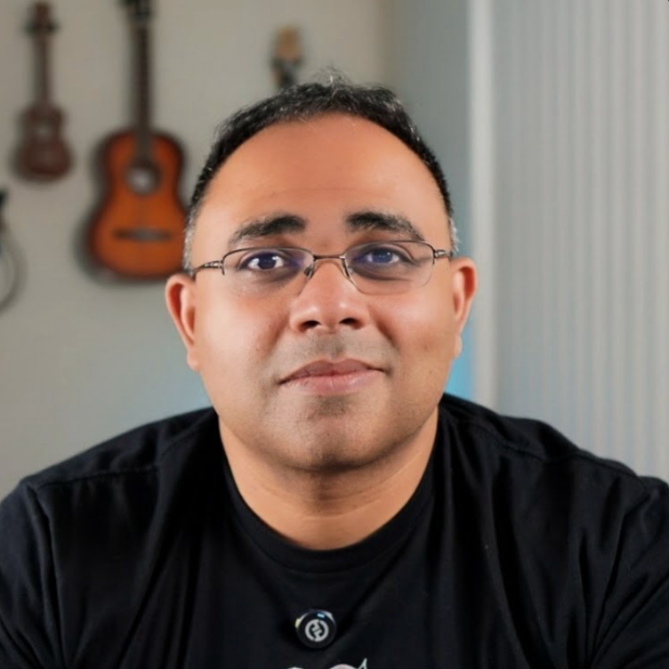 A man with glasses and short black hair, wearing a black shirt, is smiling in front of a blurred background that includes a wall with two guitars hanging on it.