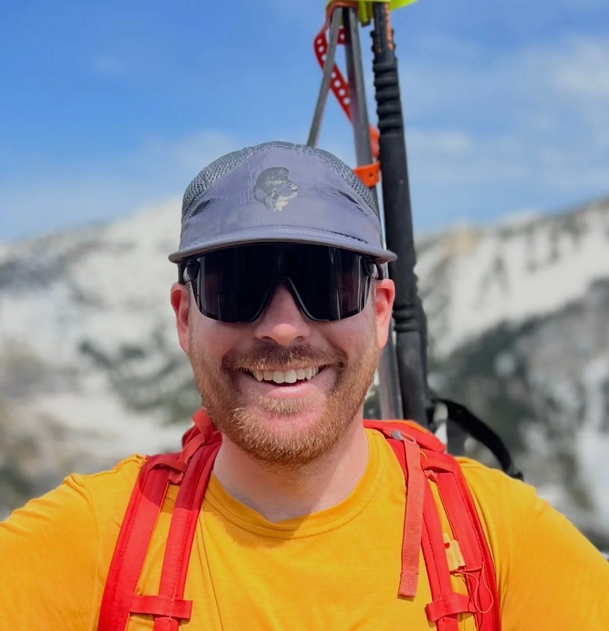 A smiling man wearing sunglasses, a gray cap with a logo, and a yellow shirt, carrying a red backpack, outdoors with a mountain background.