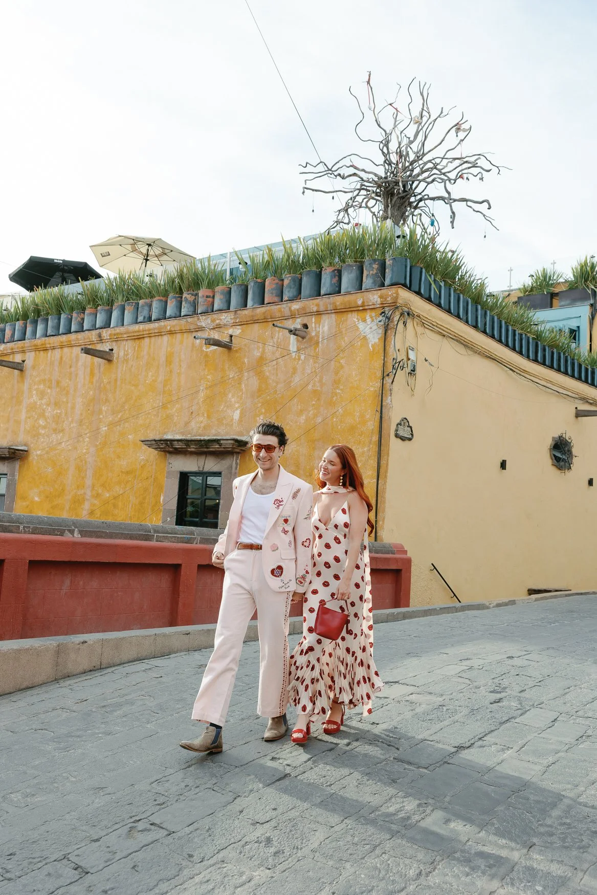 Well-dressed couple walking on a cobblestone street beside a colorful historic building in san migudel de allende with rooftop plants and umbrellas