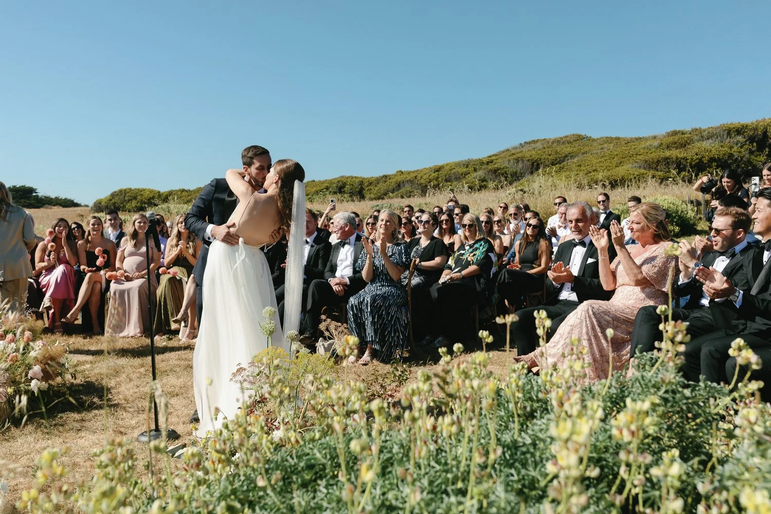 Couple getting married outdoors with audience clapping and smiling on a sunny day.