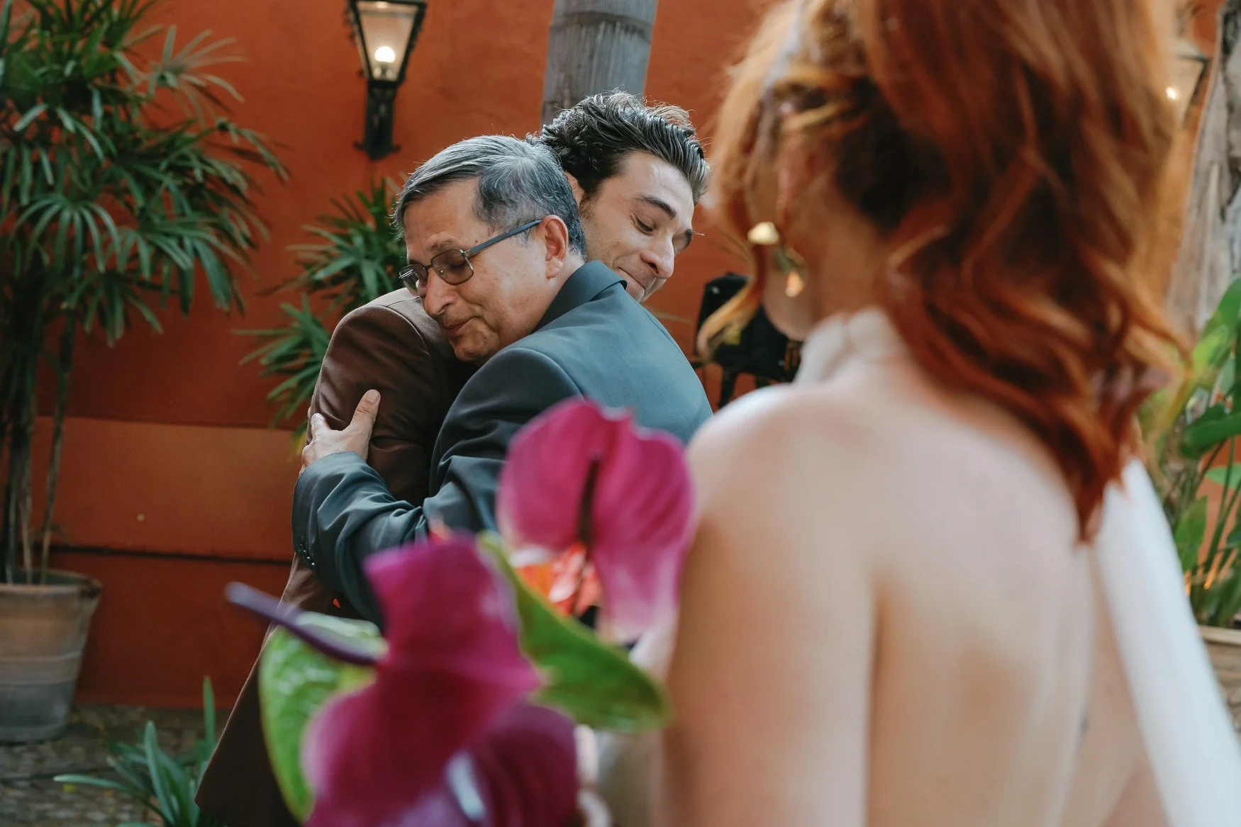 Two men embrace at a wedding reception as a bride and purple orchids appear in the foreground.