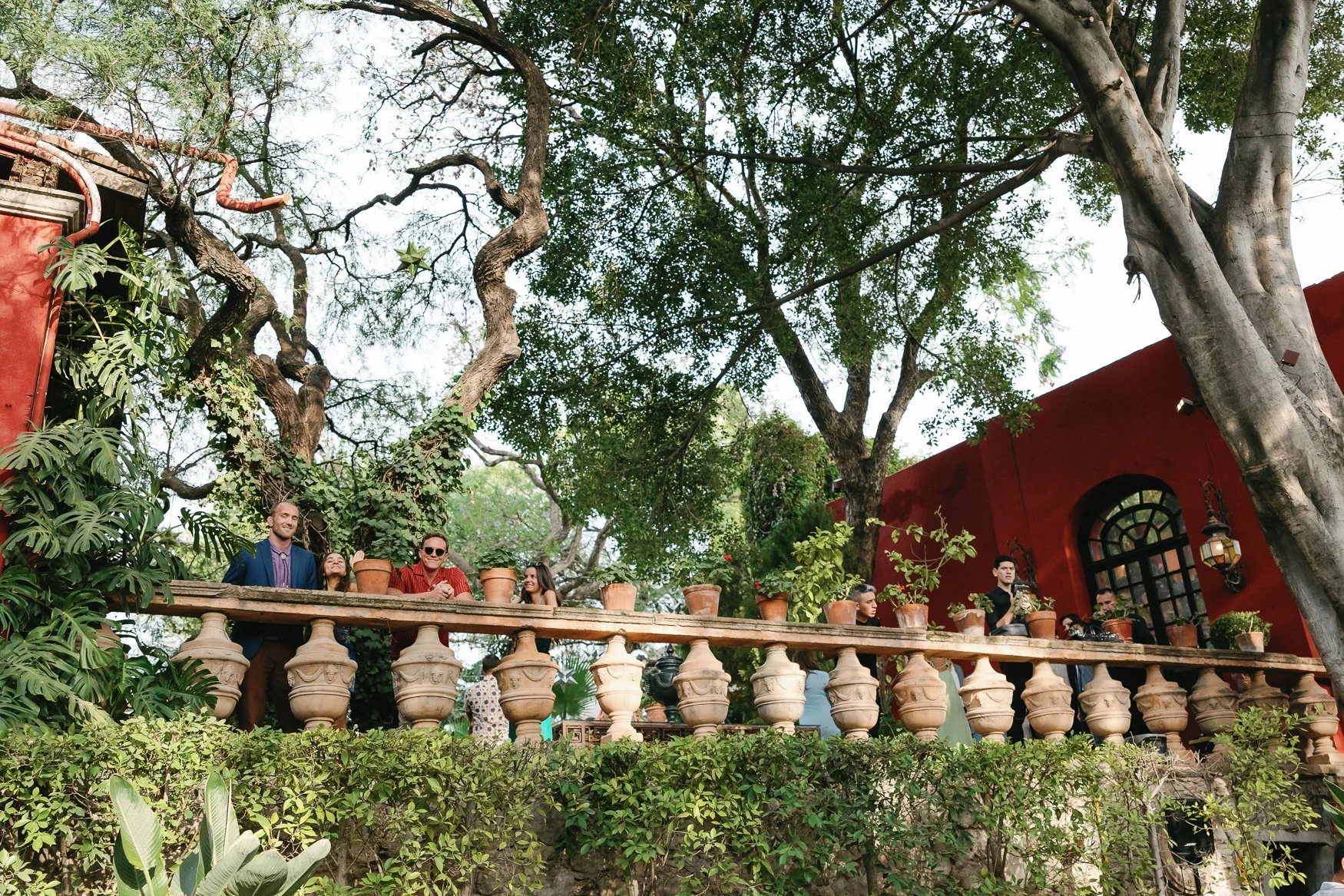 People gathered on a lush, tree-shaded terrace with terracotta potted plants, ornate stone balustrades, and a red building.